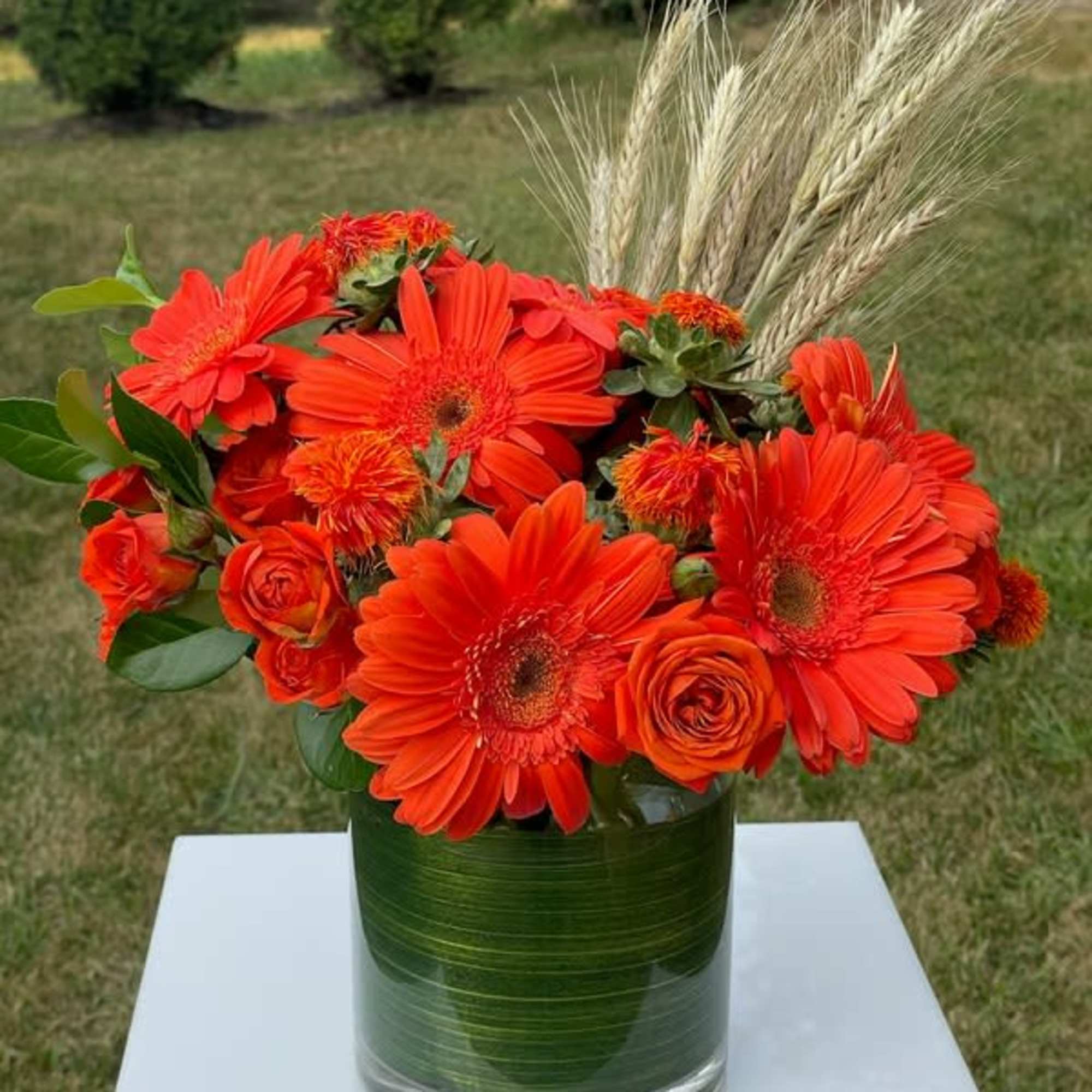 Orange gerbera daisies and roses in a glass vase with wheat stems