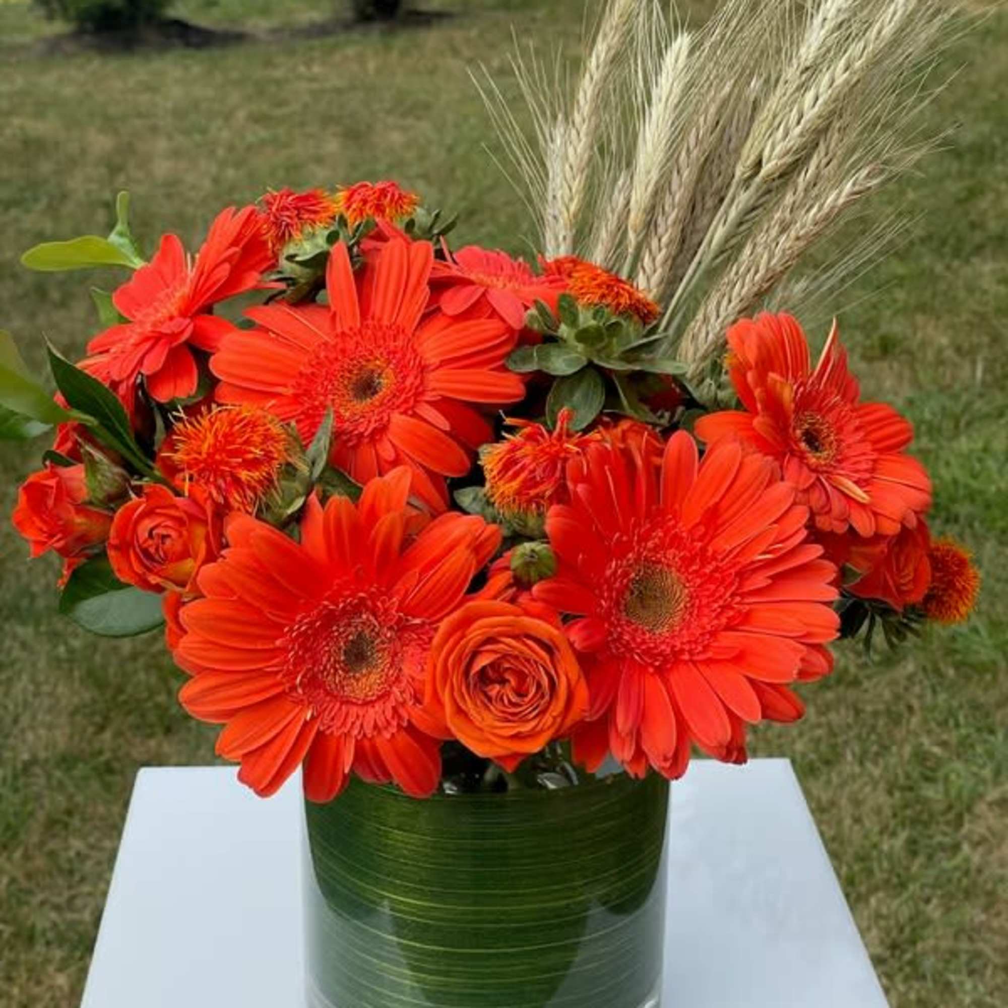 Orange gerbera daisies and roses in a glass vase with wheat stems