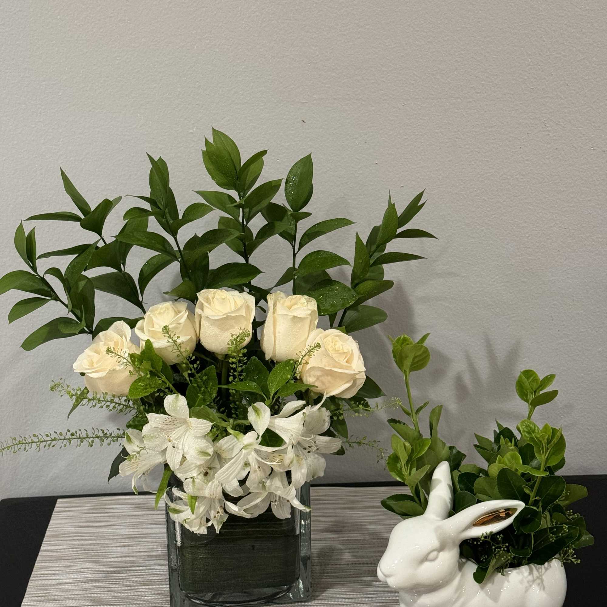 White roses and white alstroemeria in a glass vase beside a bunny planter