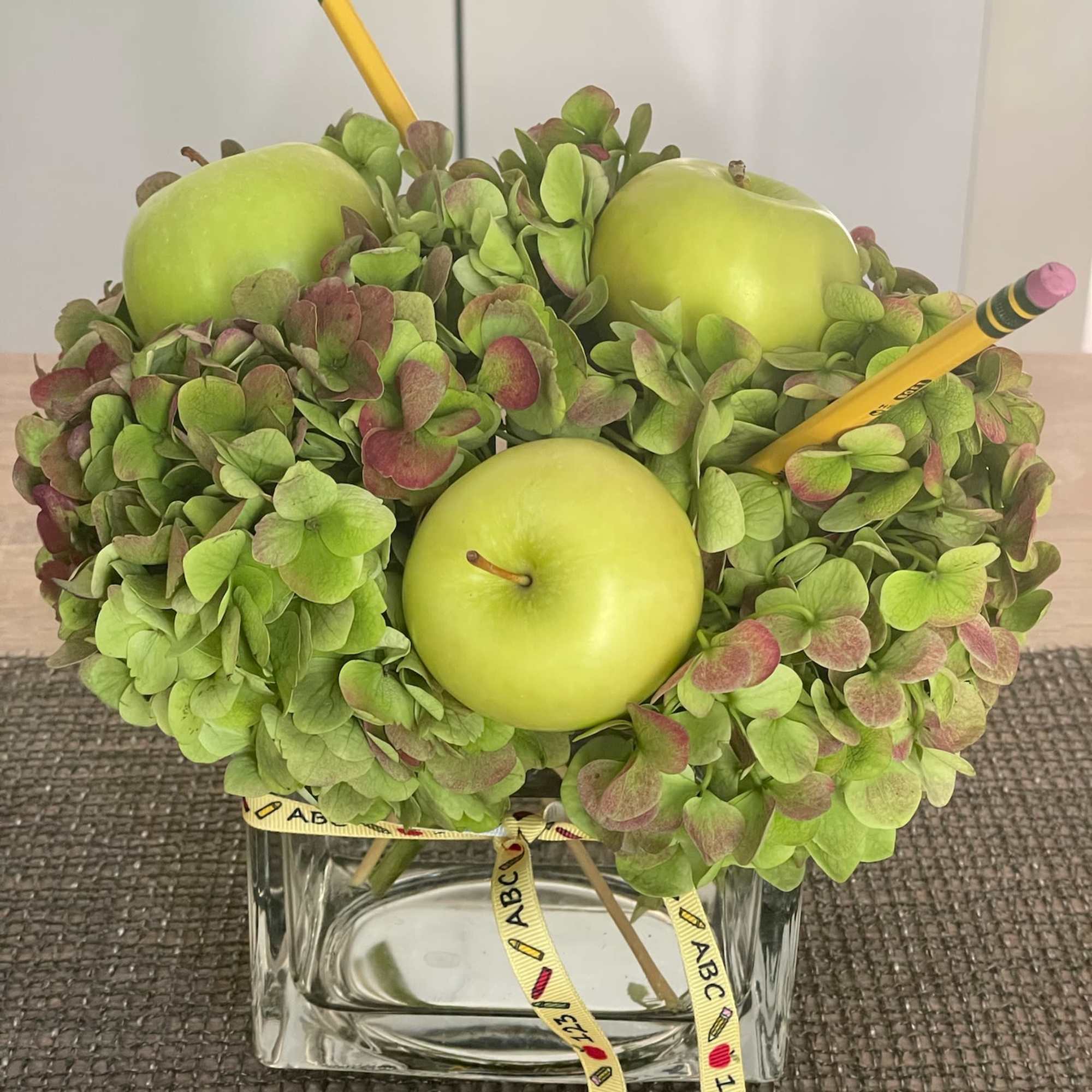 Green hydrangea arrangement with apples and pencils in a glass vase
