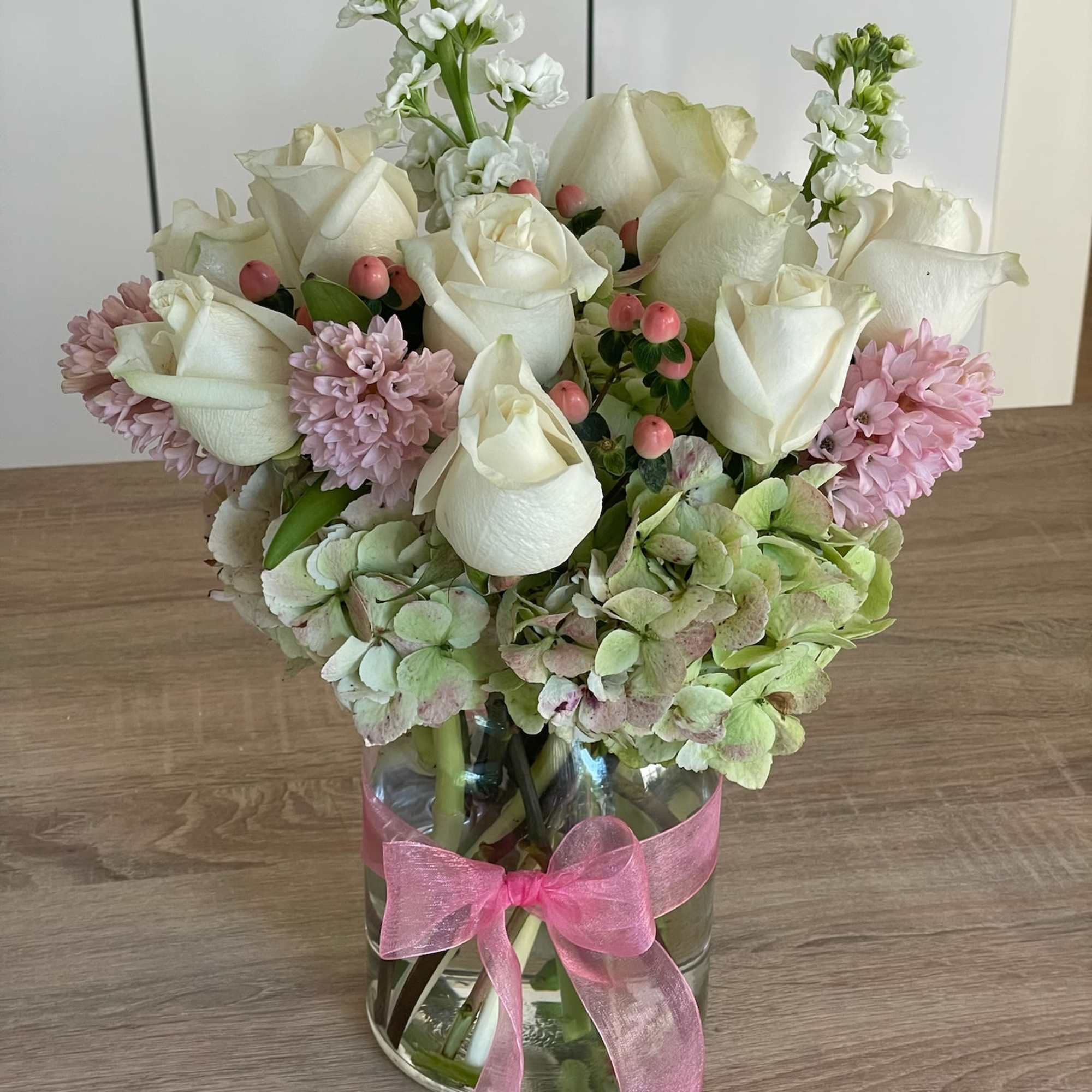 White roses and pink flowers arranged in a glass vase with a pink ribbon