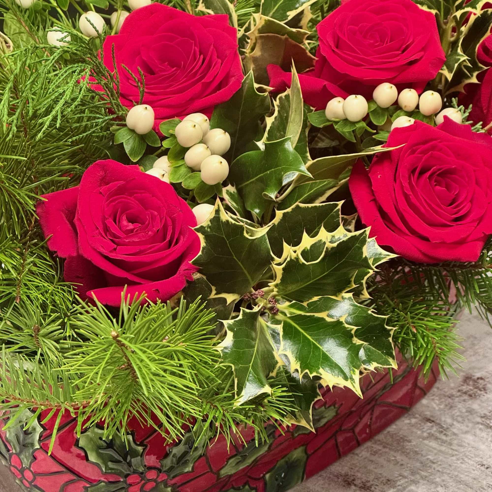 Red roses with holly and white berries in a red glass container
