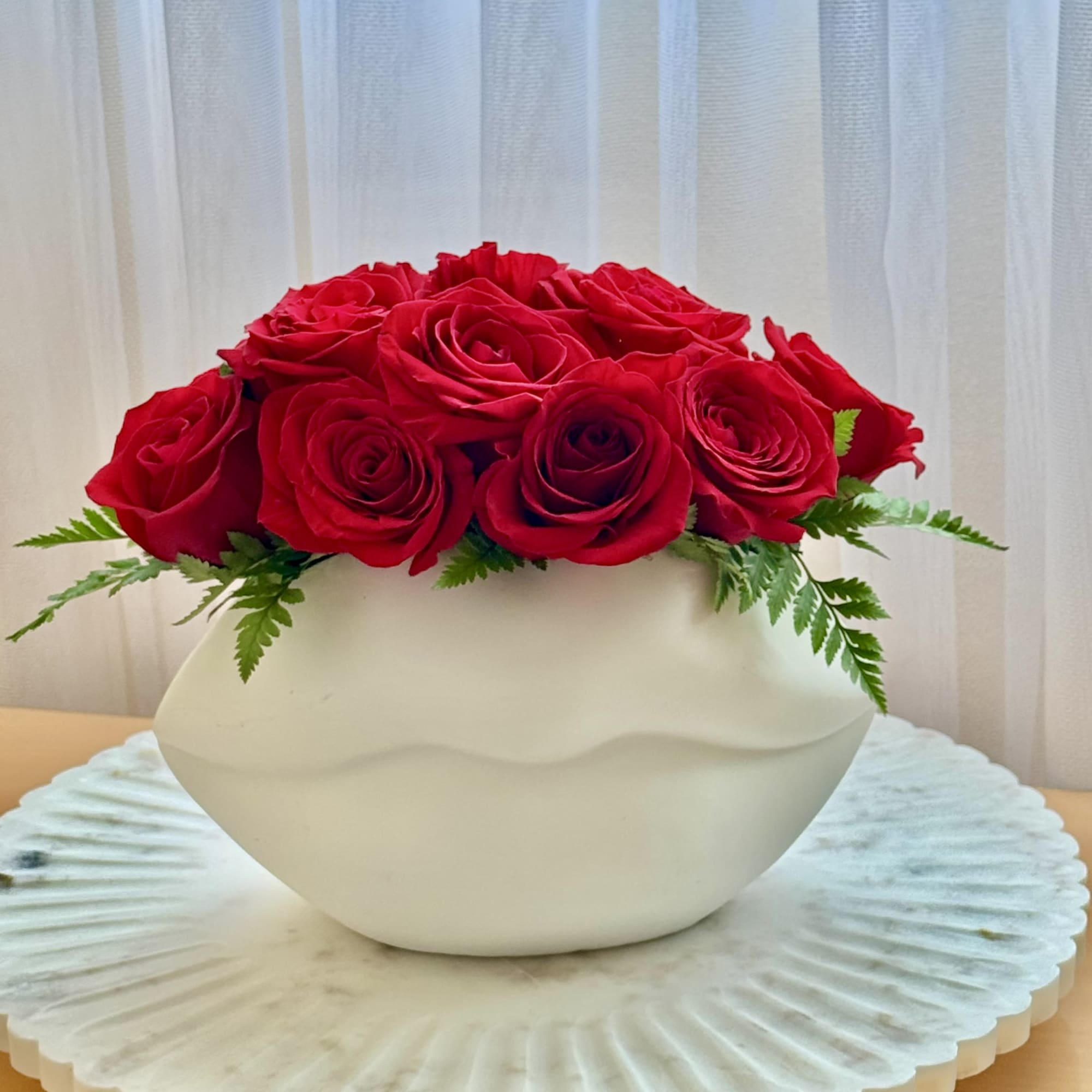 Red roses arranged in a white bowl vase with fern accents