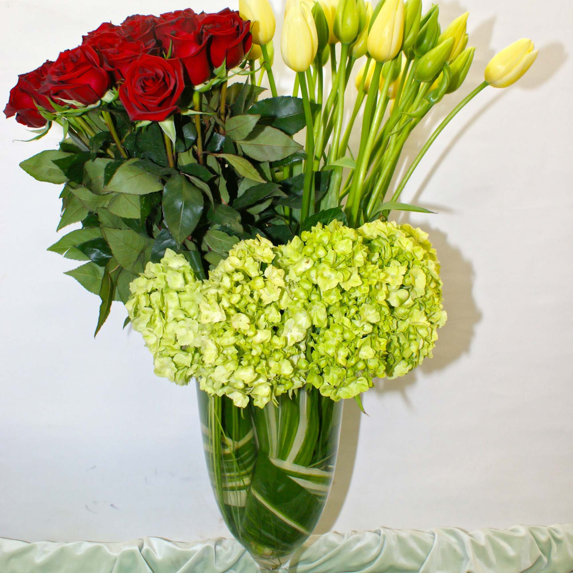 Red roses and yellow tulips arranged in a clear glass vase with green hydrangeas