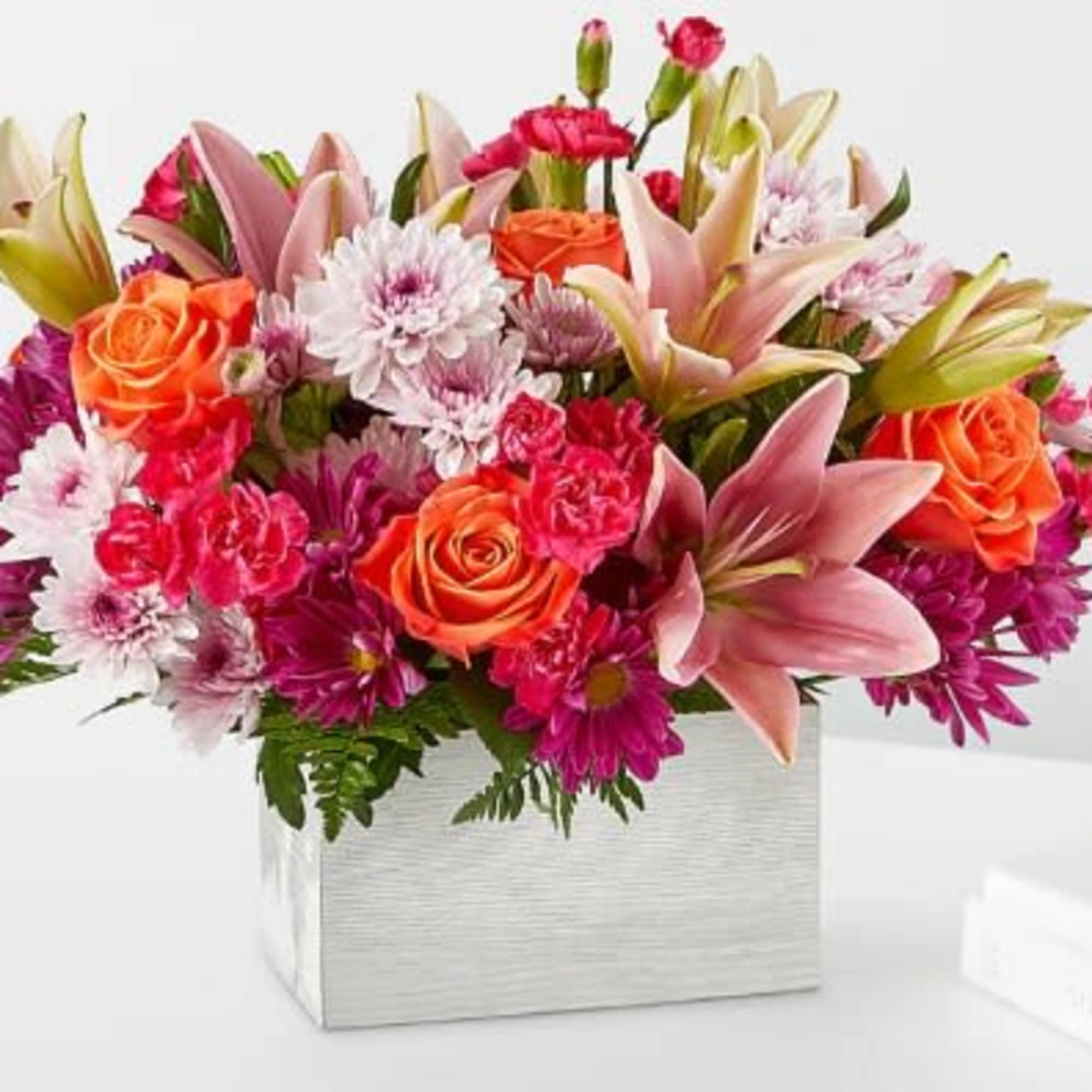 Bright arrangement of pink lilies, orange roses, and mixed blooms in a white rectangular container