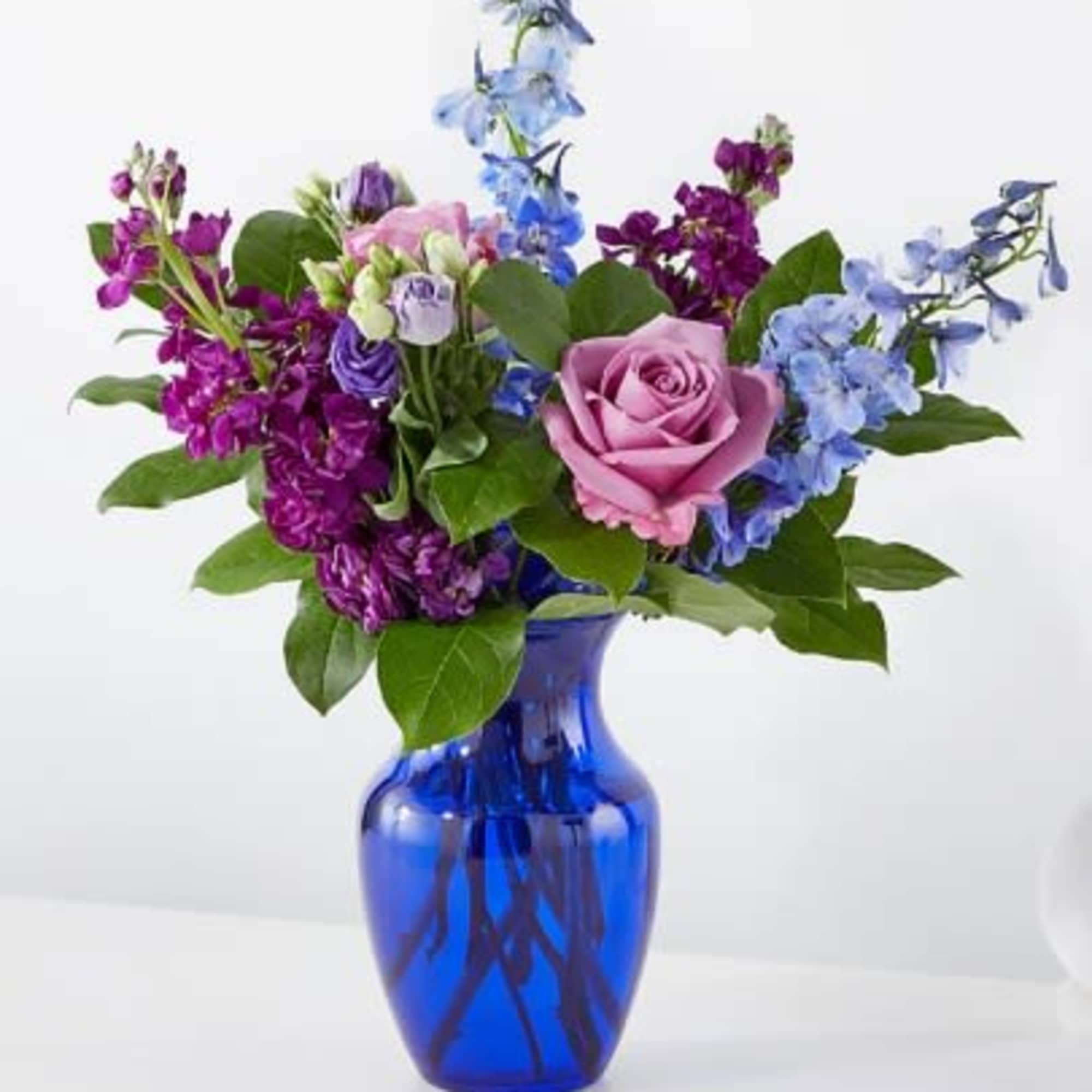 Purple, blue, and pink flower arrangement with roses and delphinium in a cobalt blue glass vase