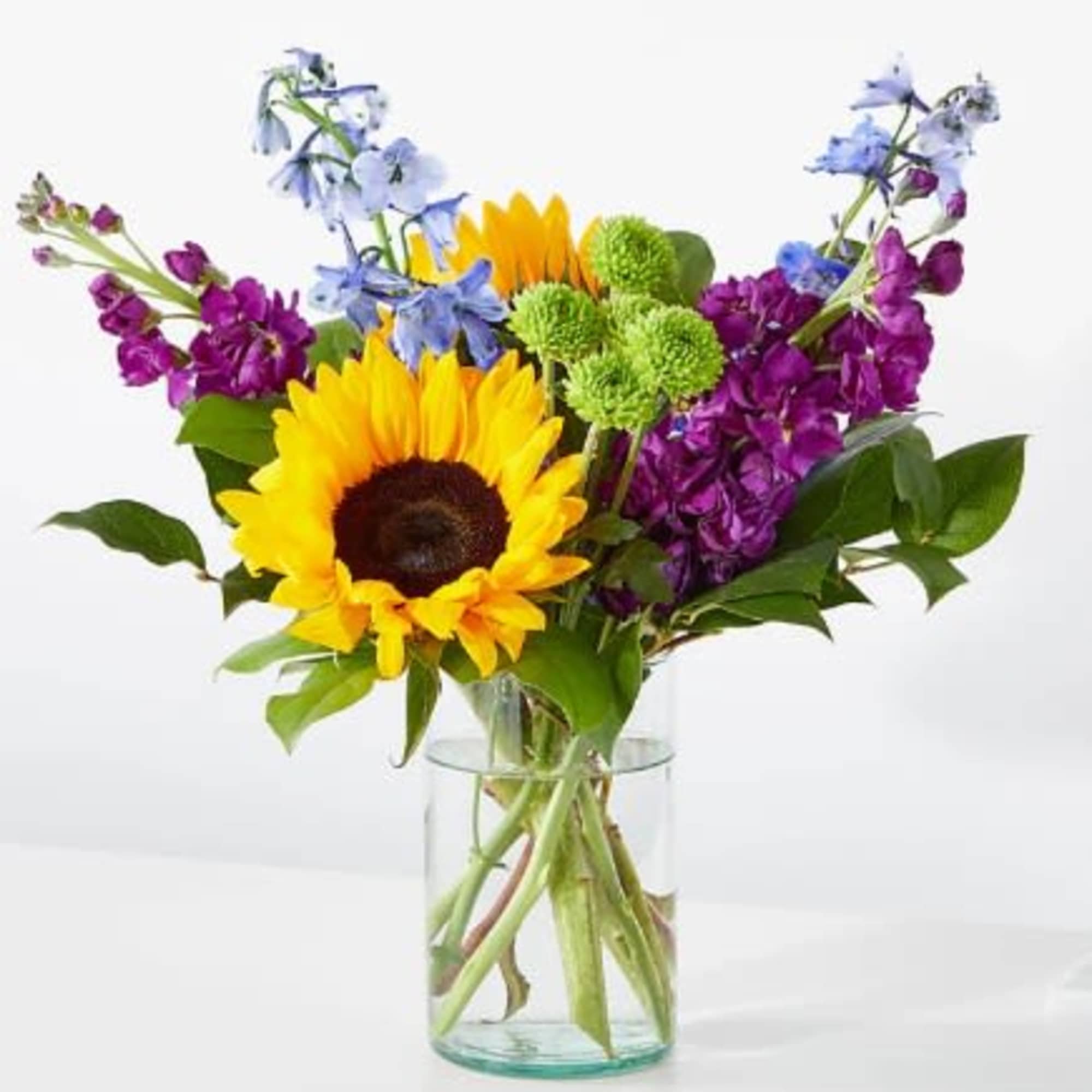 Bright sunflower, purple stock, blue delphinium, and green mums in a clear glass vase.