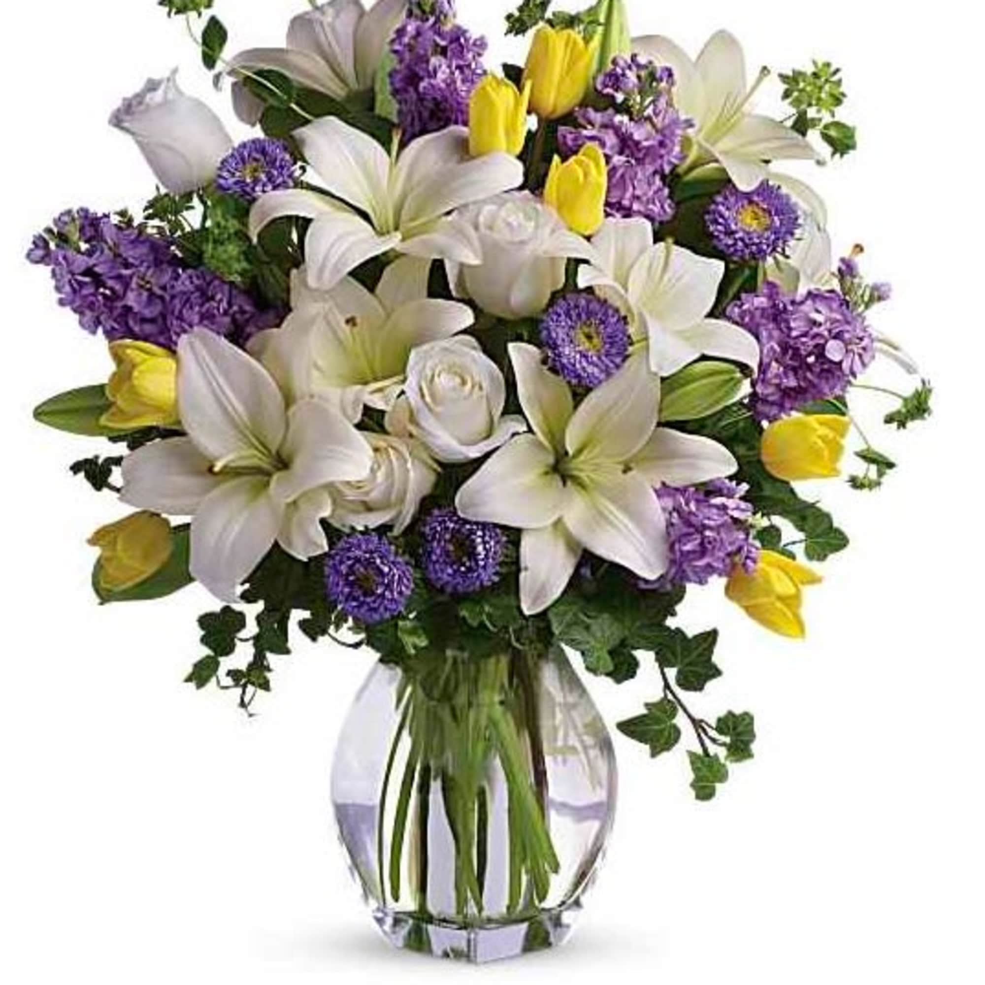 White lilies and purple flowers arranged in a glass vase with yellow tulips