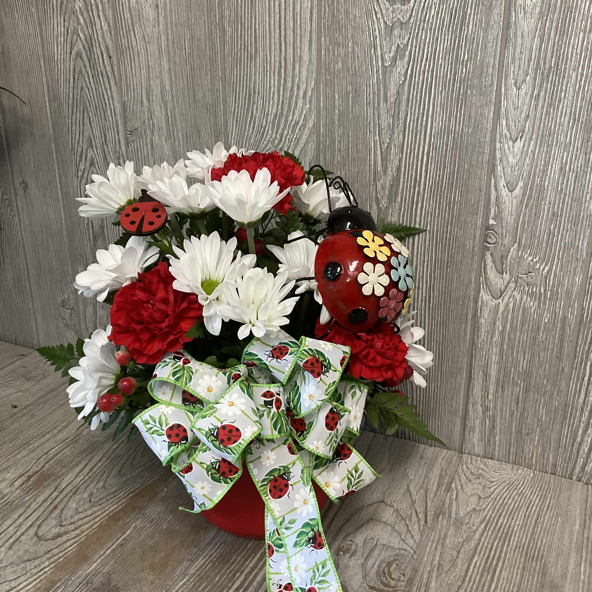 Red and white flower arrangement in a red pot with ladybug decorations and ribbon