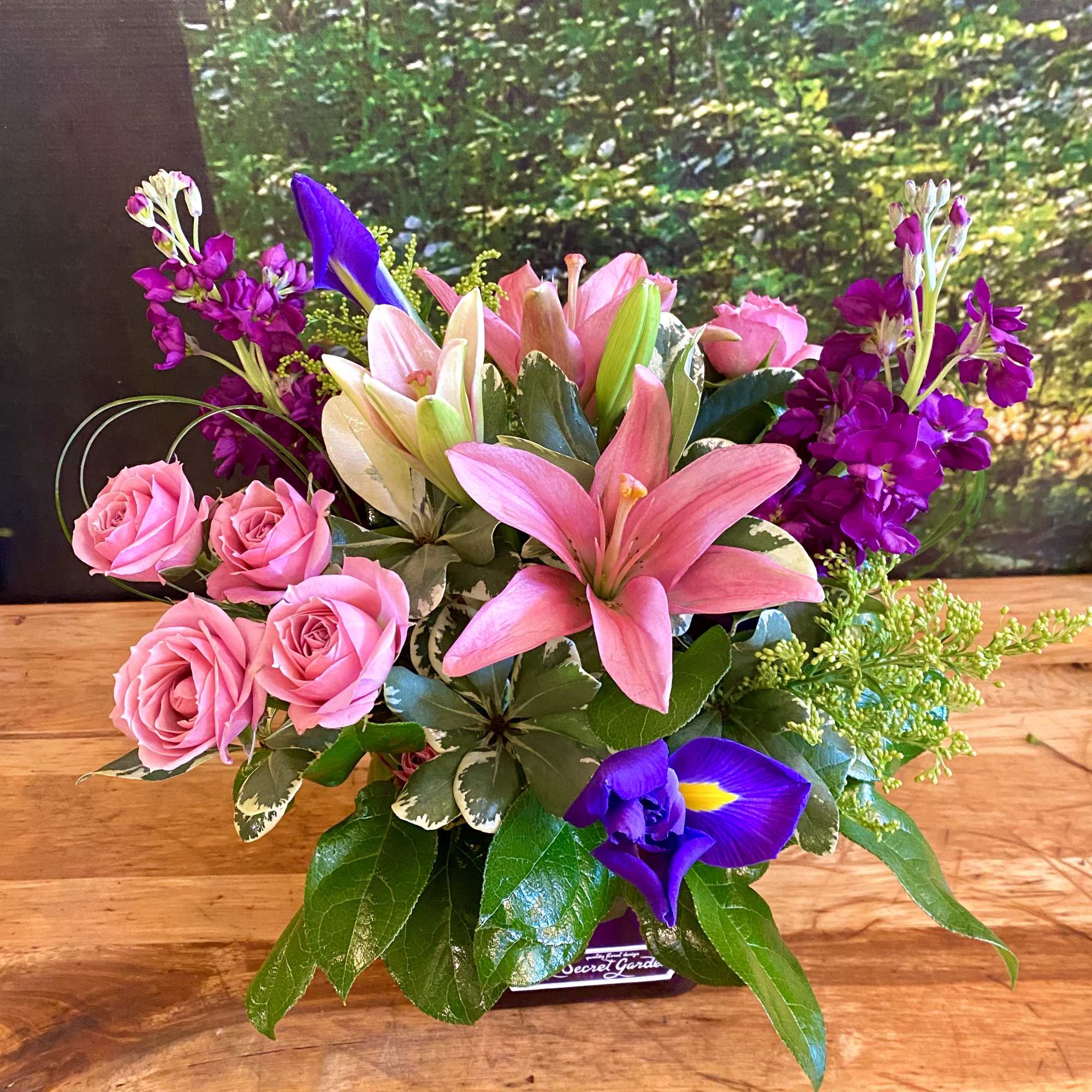Pink lilies and roses arranged with purple flowers in a low container