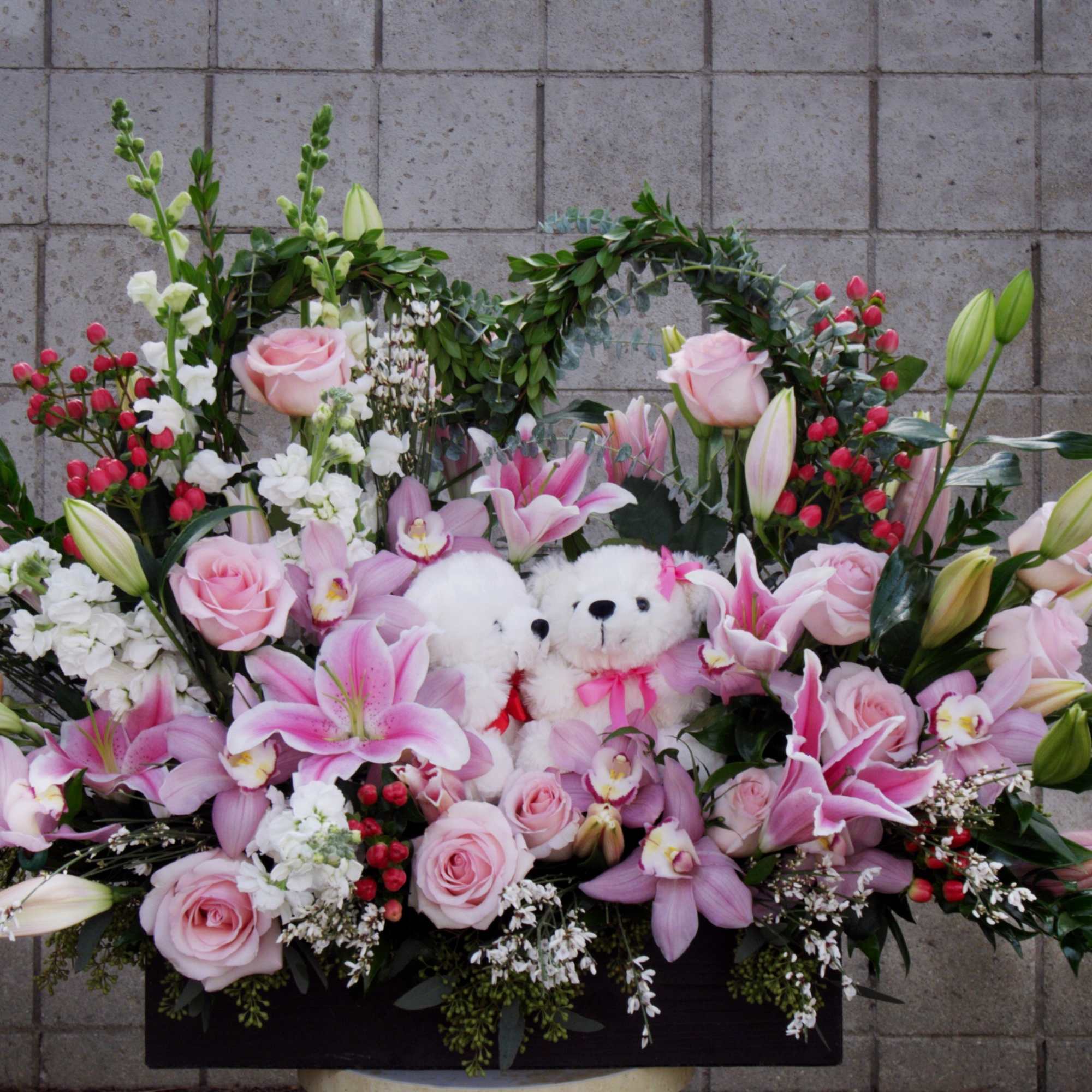 Pink lilies and roses arranged around two white teddy bears in a heart-shaped display.