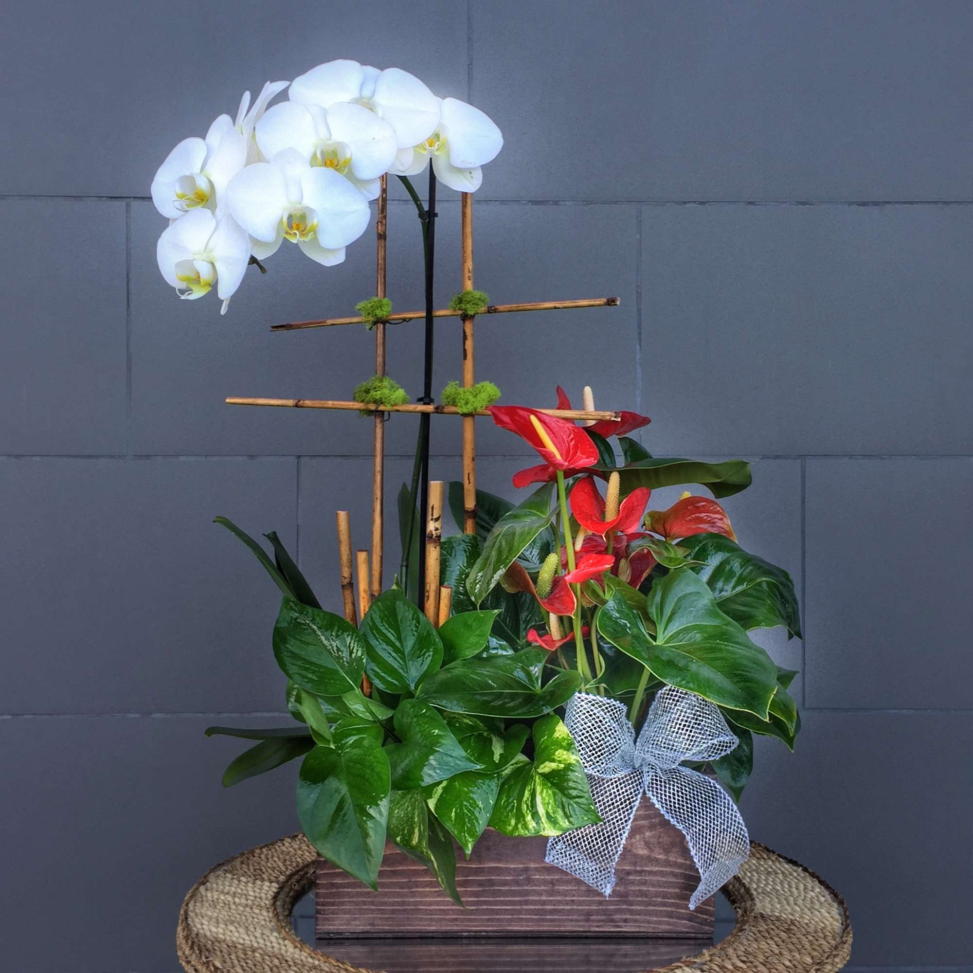 White orchids and red anthuriums in a wooden planter with a ribbon bow