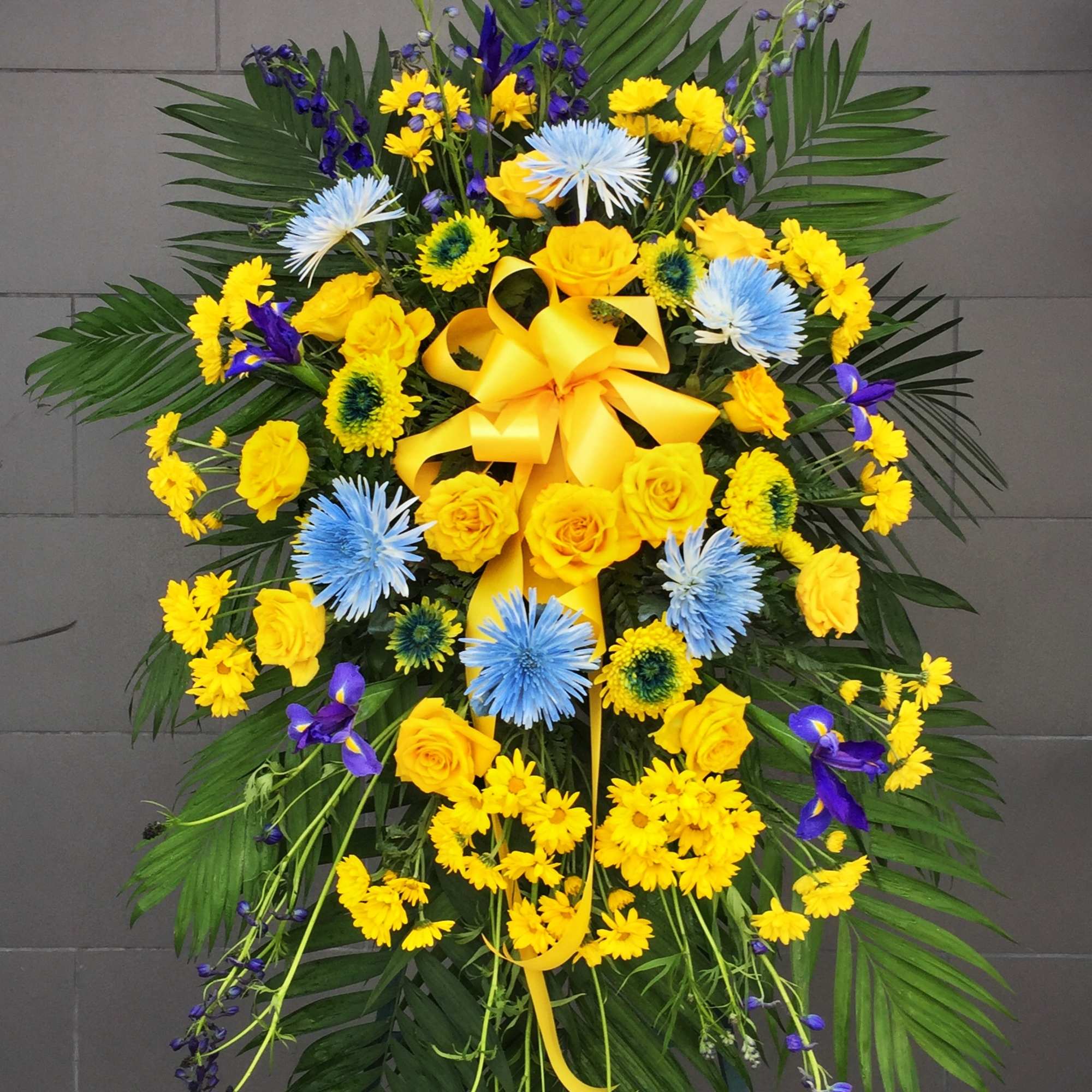 Standing floral spray with yellow roses, blue flowers, and a yellow ribbon