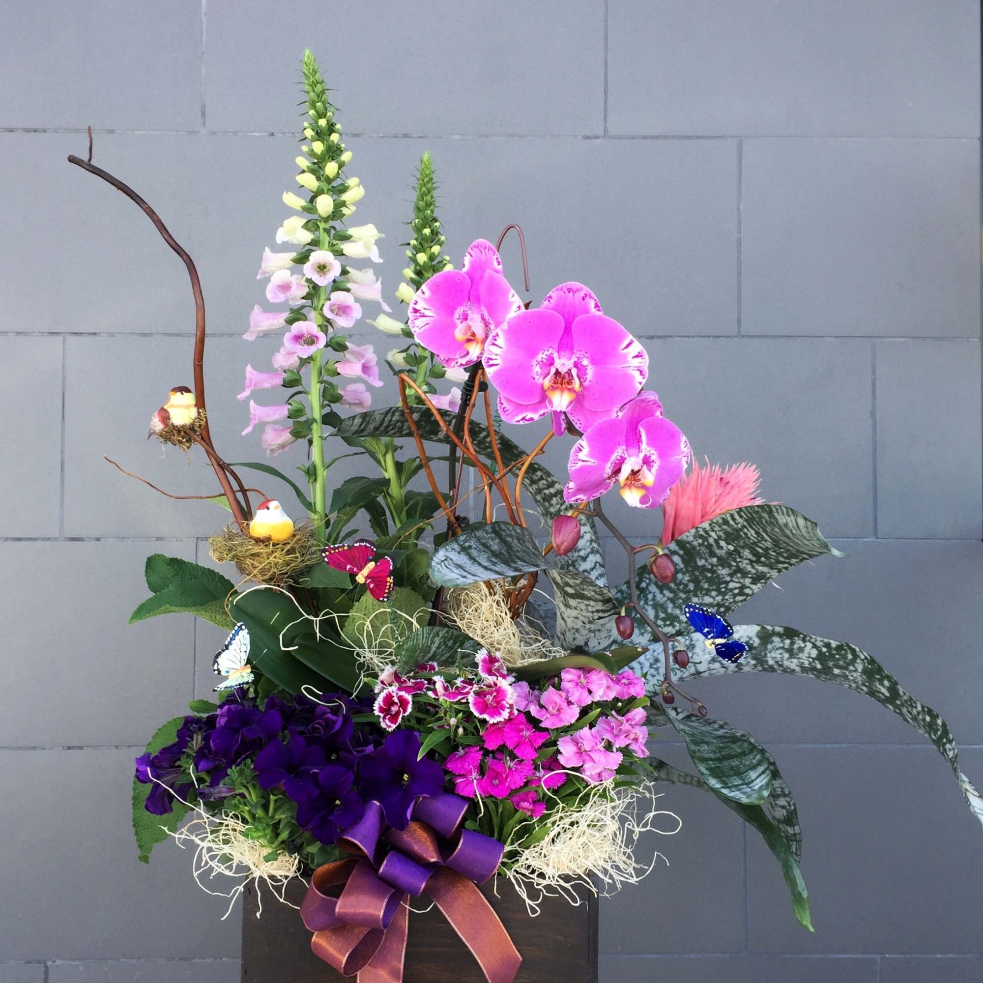 Tall floral arrangement with pink orchids, purple blooms, and a ribboned box base