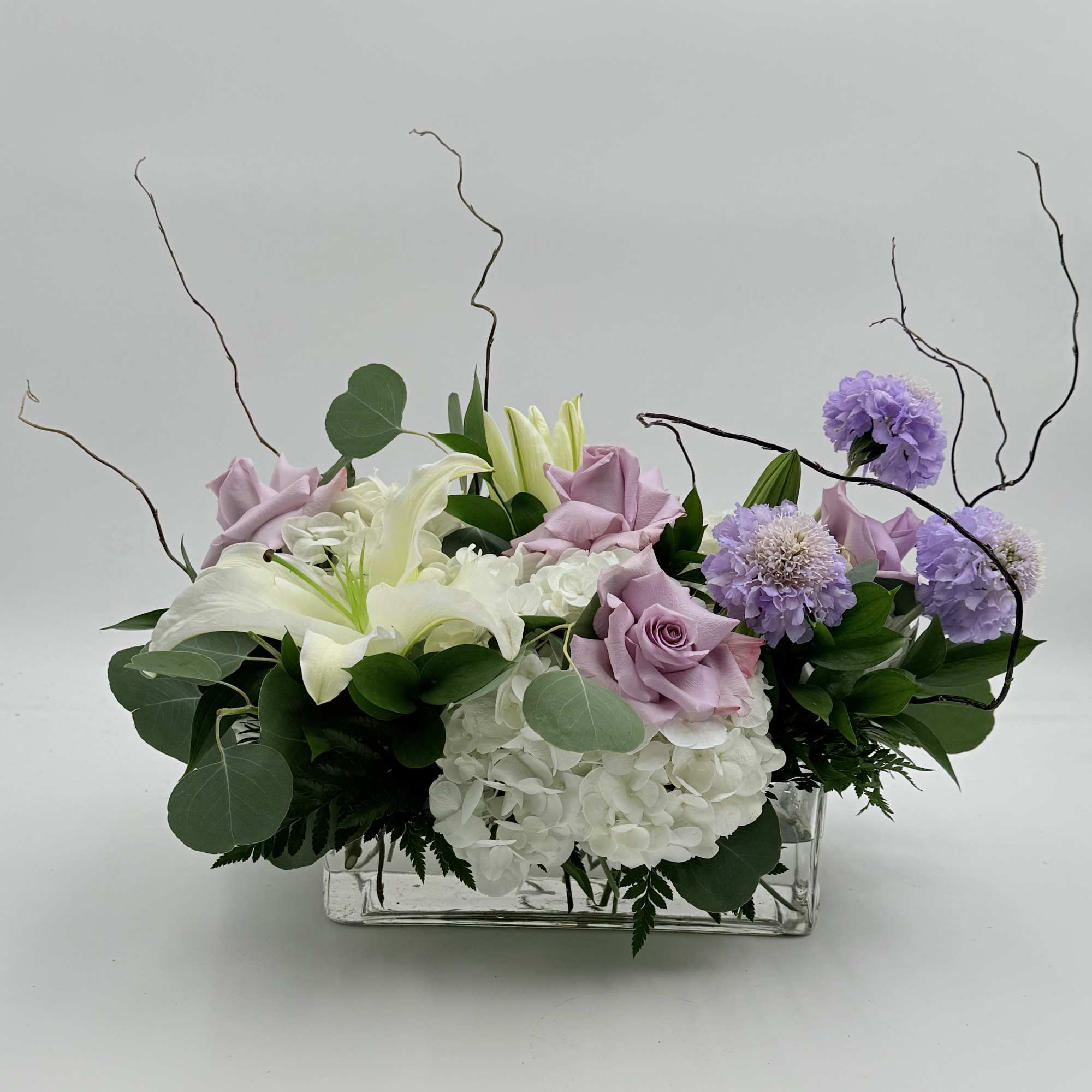 Lavender roses and white lilies arranged with purple blooms in a clear rectangular vase.