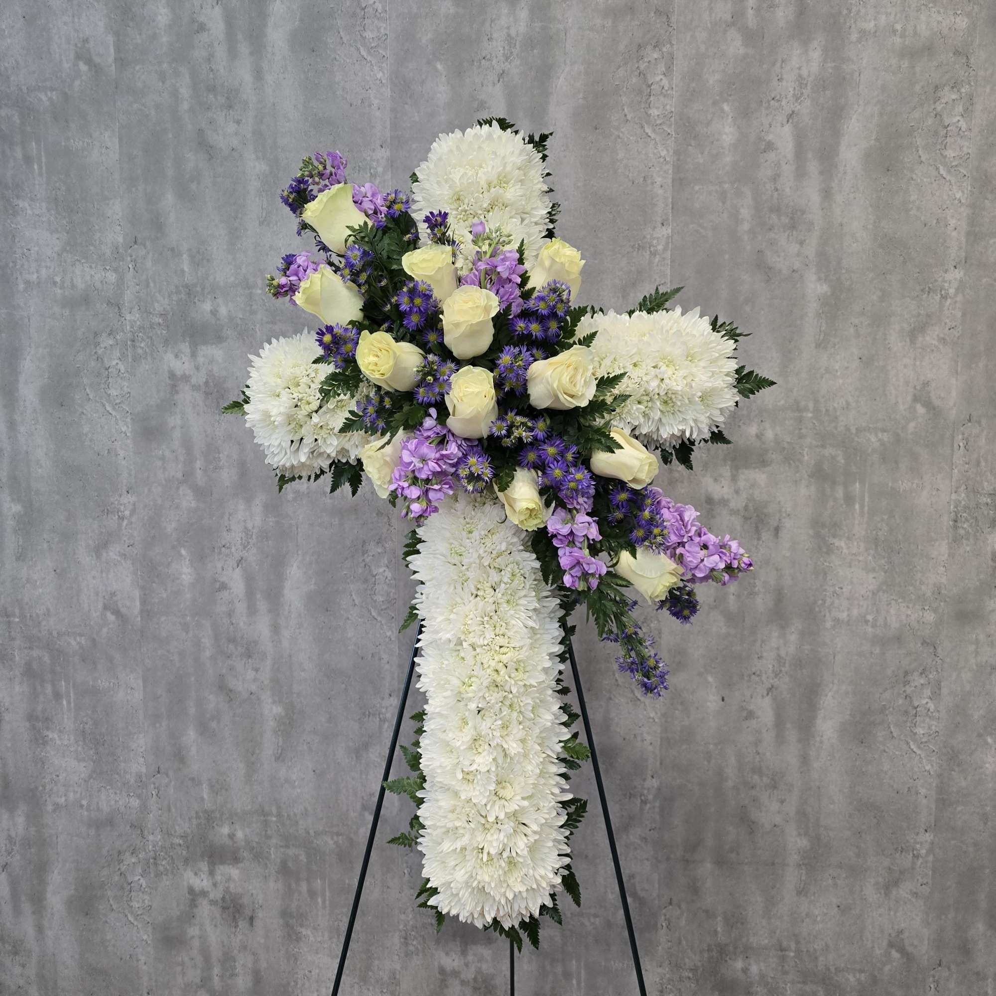 White floral cross arrangement with pale yellow roses and purple accents on a stand