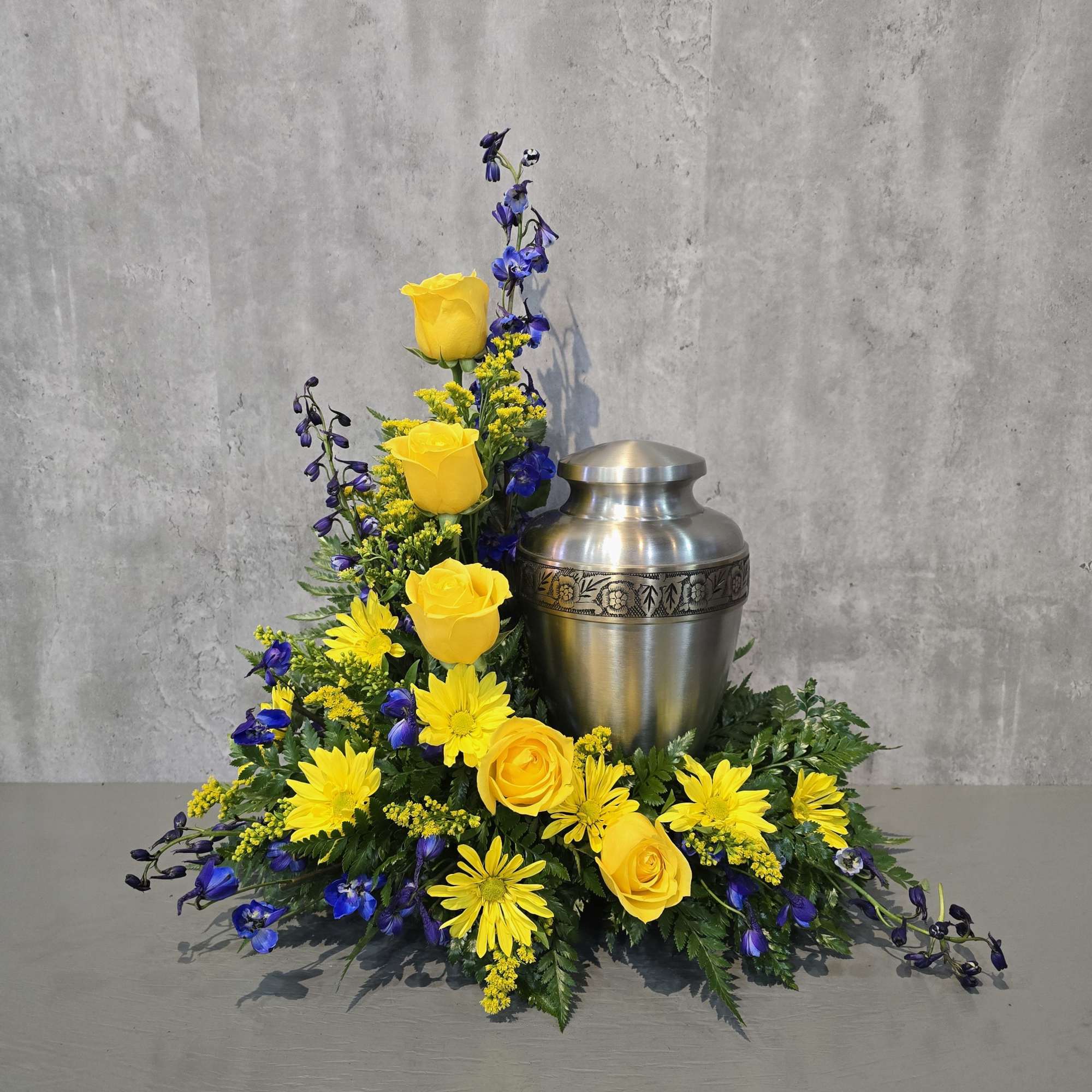 Yellow roses and daisies arranged around a silver urn with purple accents