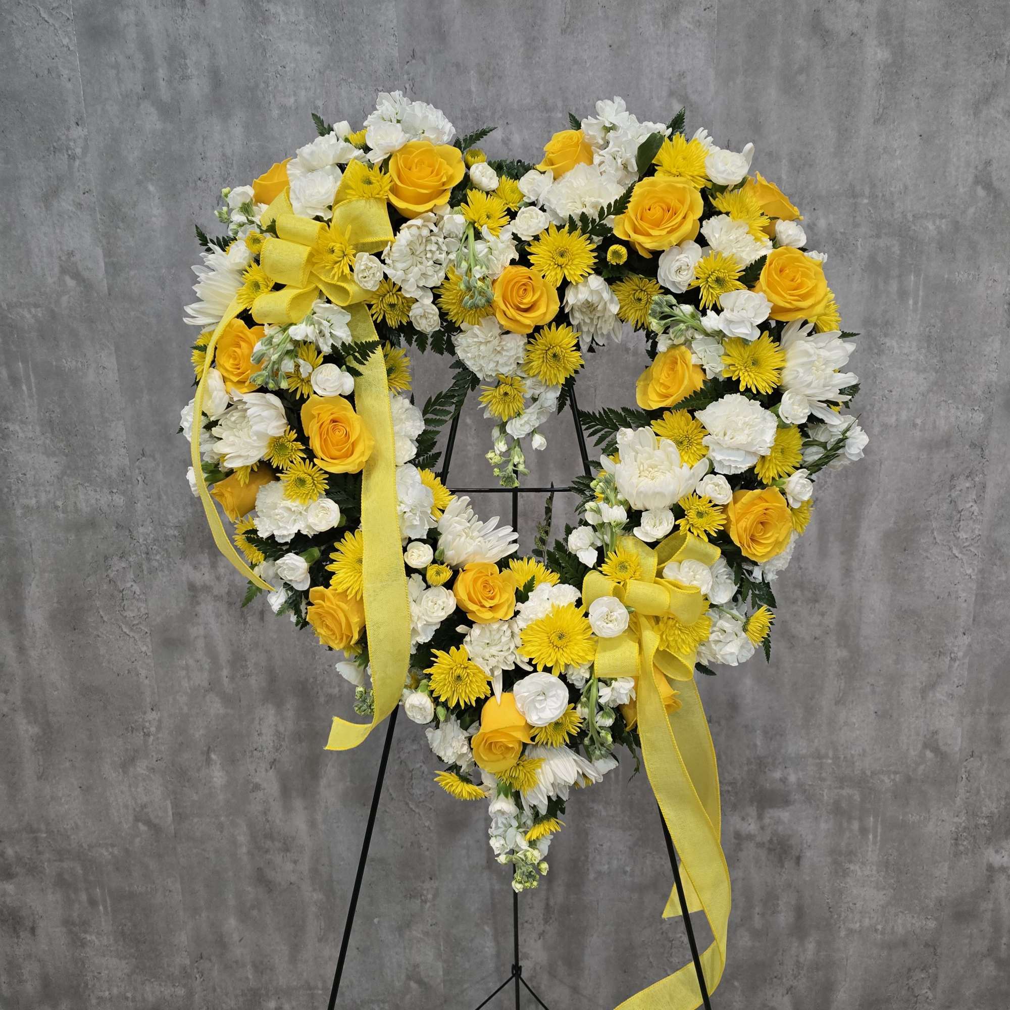 Heart-shaped floral wreath with yellow and white flowers on a black stand