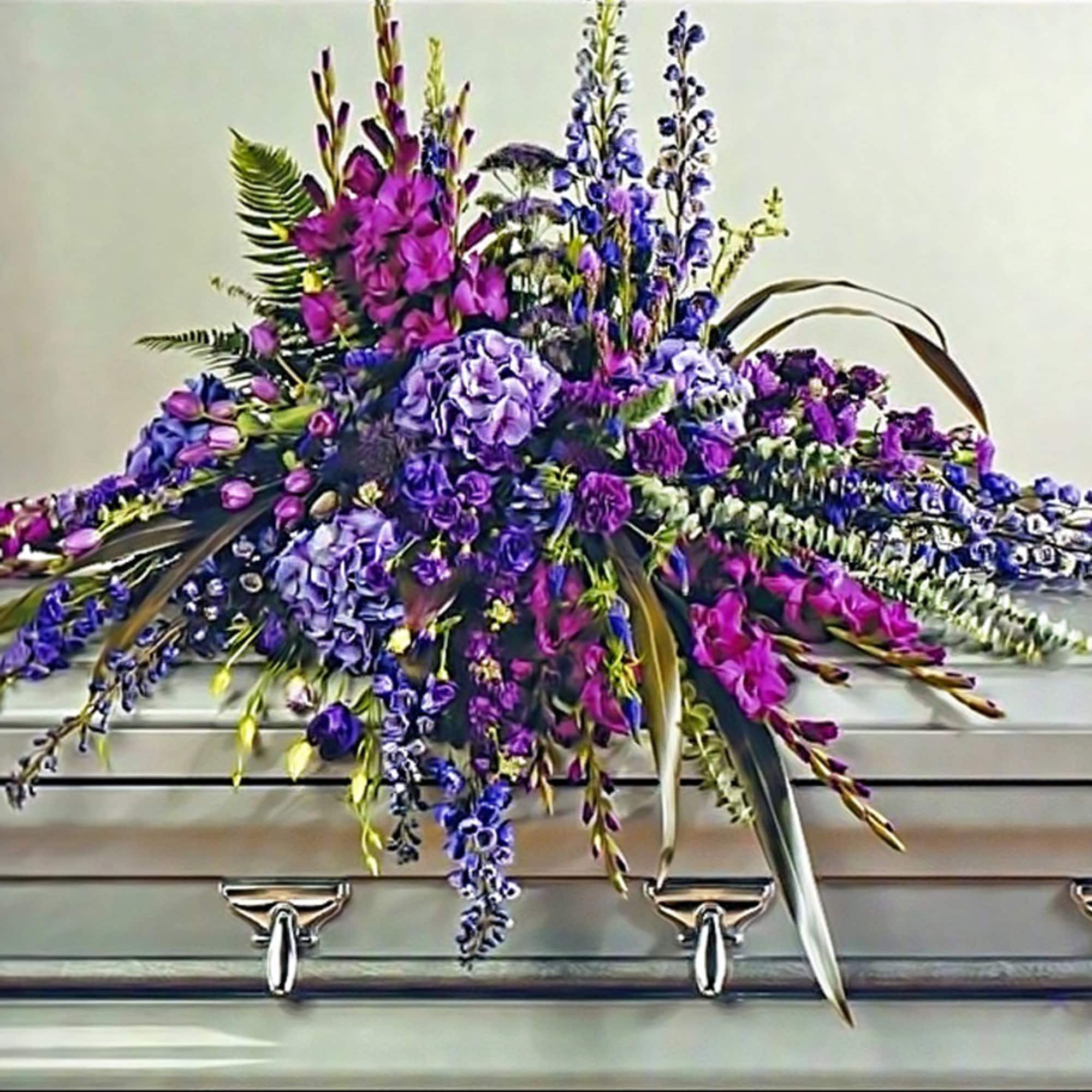 Purple and lavender funeral casket spray with mixed flowers cascading over a silver casket