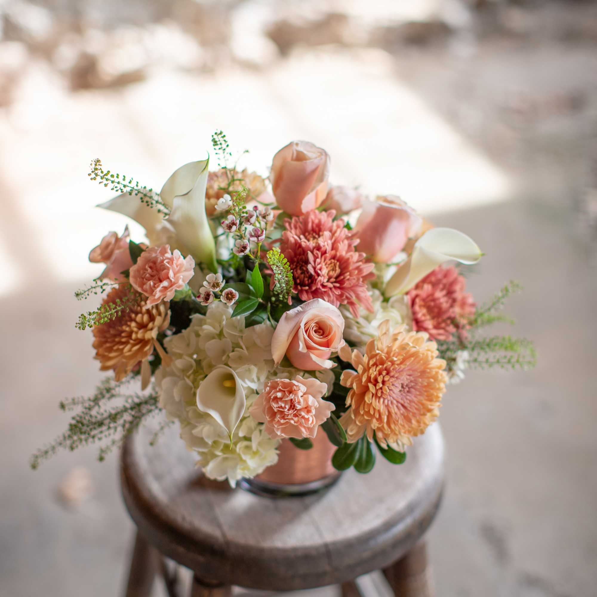 Peach and white floral bouquet in a copper vase on a wooden stool