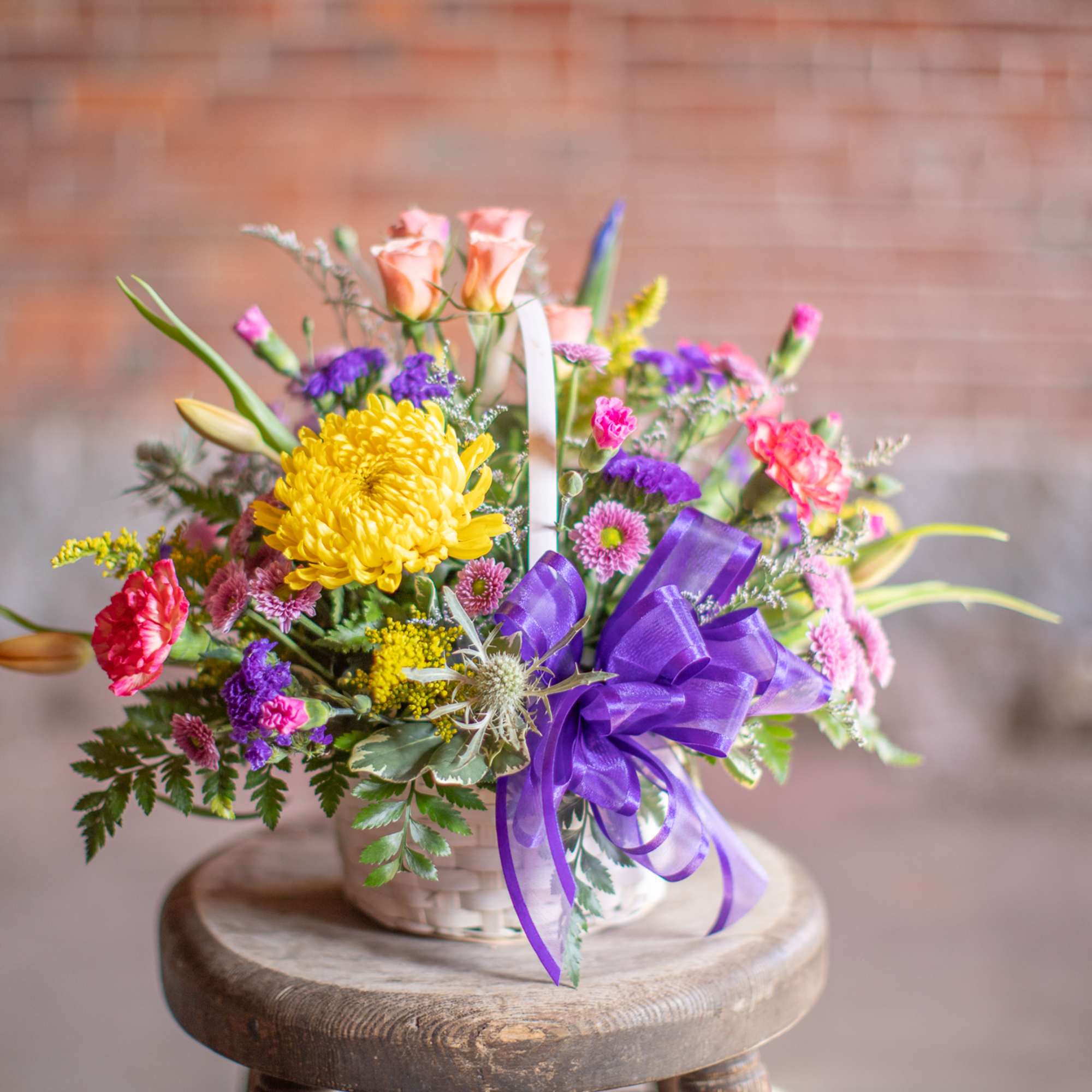 Colorful mixed flower arrangement in a white basket with a purple ribbon