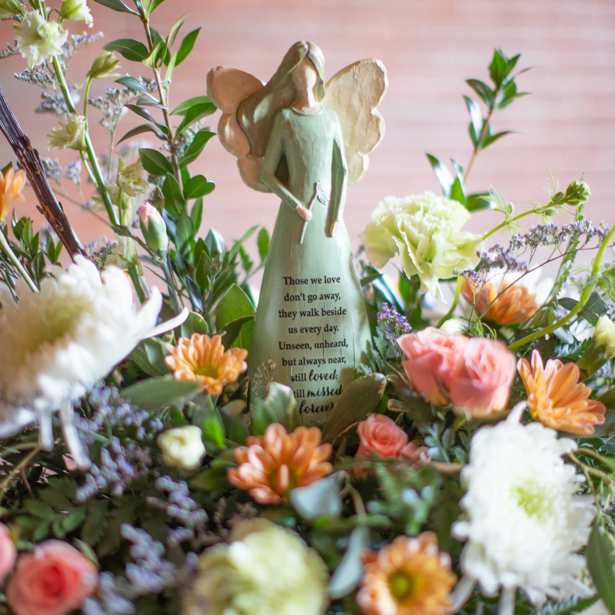 Angel figurine with inscription surrounded by peach, white, and pink flowers in a low round arrangement