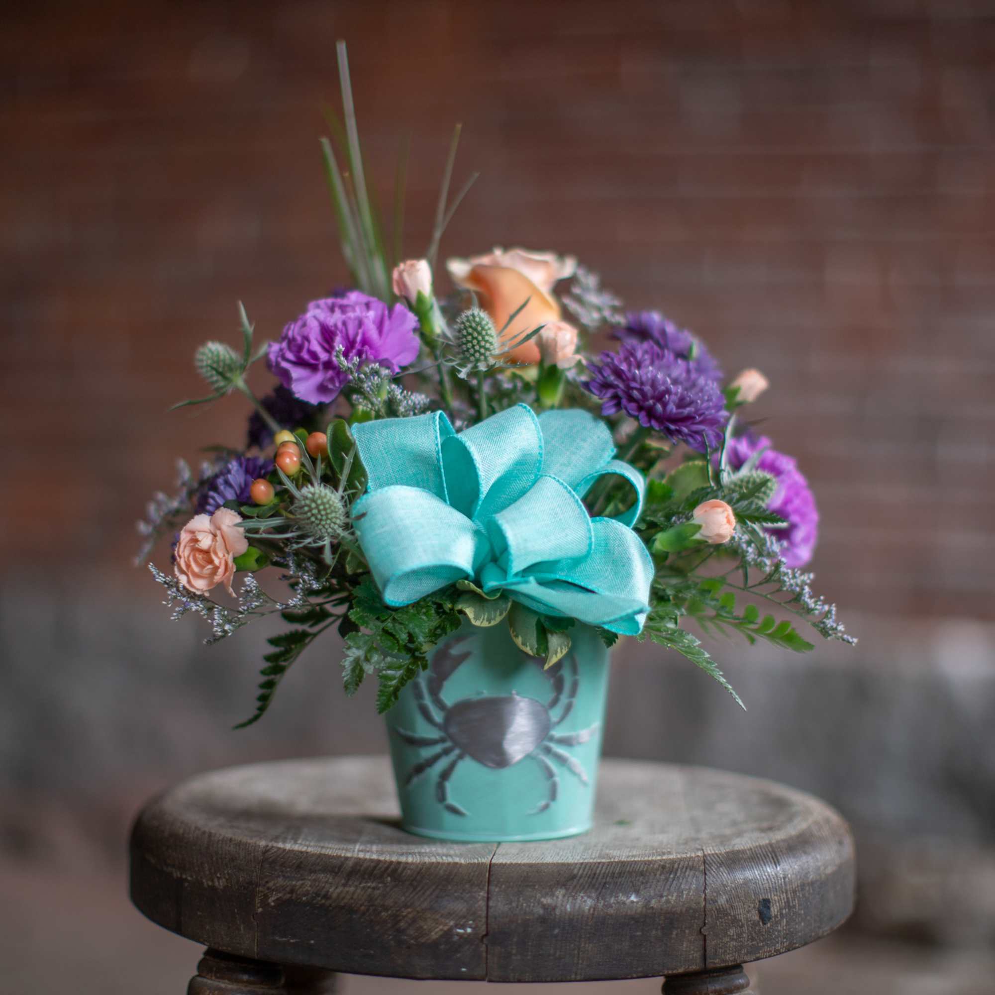 Purple and peach flower arrangement in a turquoise pot with a large bow