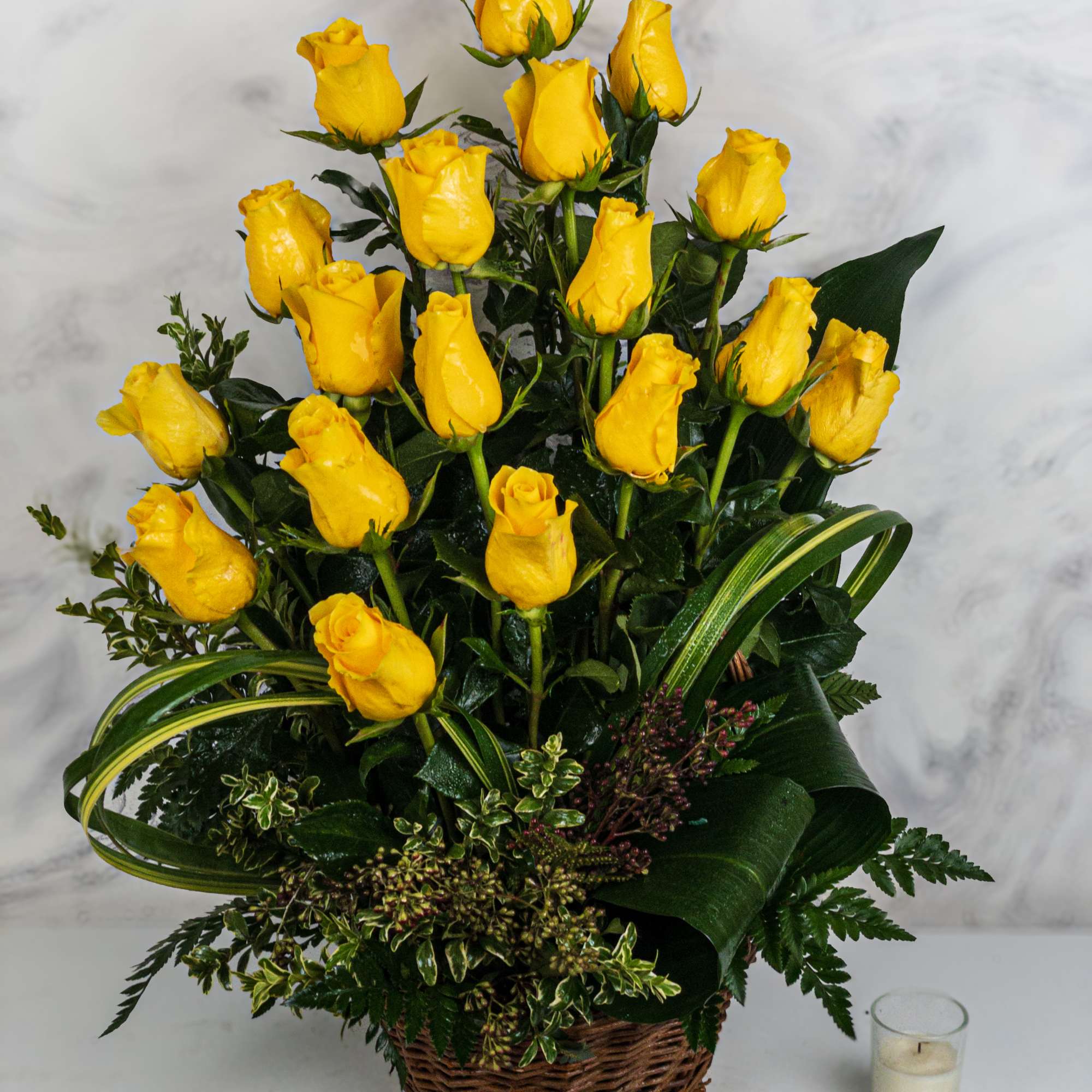 Basket arrangement of yellow roses with greenery and a small candle beside it