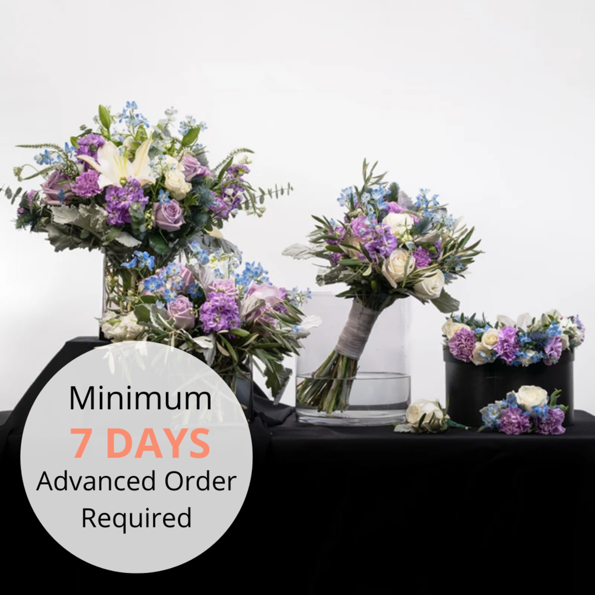 Coordinated purple, white, and blue floral bouquets and arrangements on a black cloth with glass and black vases.