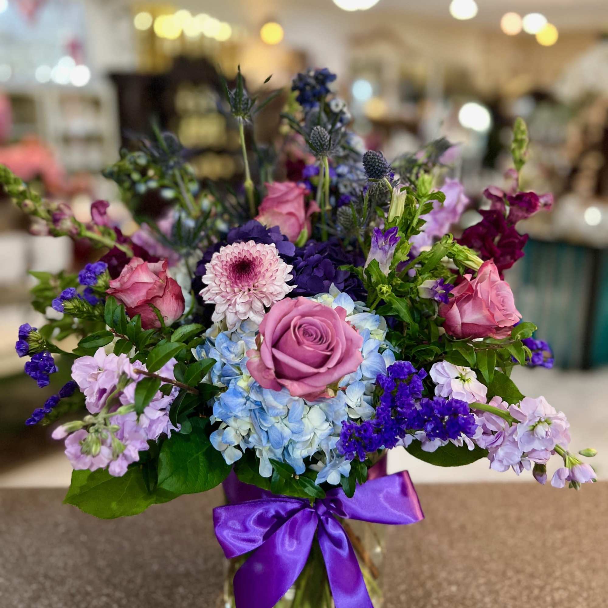 Mixed pink, purple, and blue flowers arranged in a glass vase with a purple ribbon.