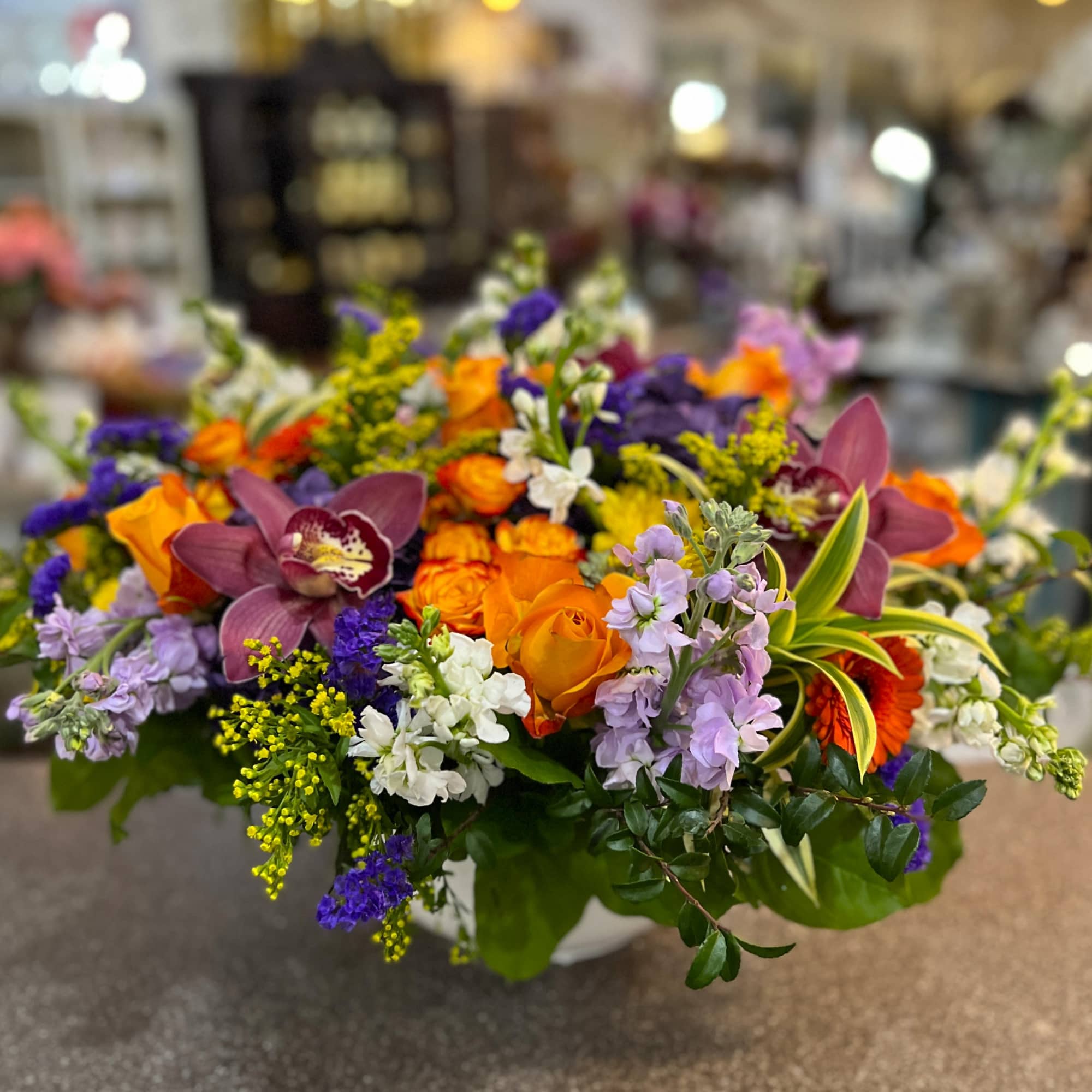 Mixed bouquet of orange roses, purple orchids, and lavender flowers in a low container