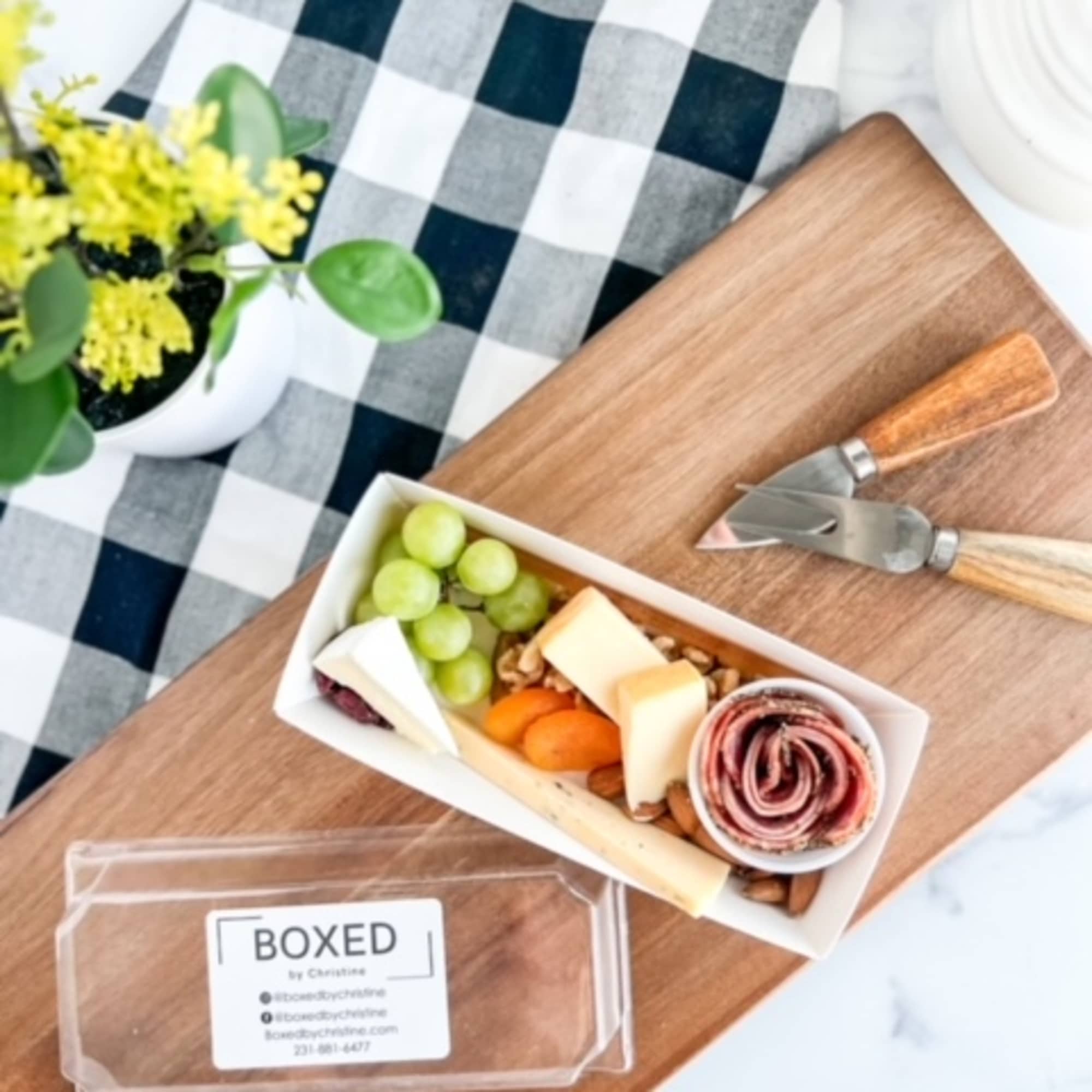 Boxed snack tray with grapes, cheese, nuts, and rolled meat beside a small potted plant