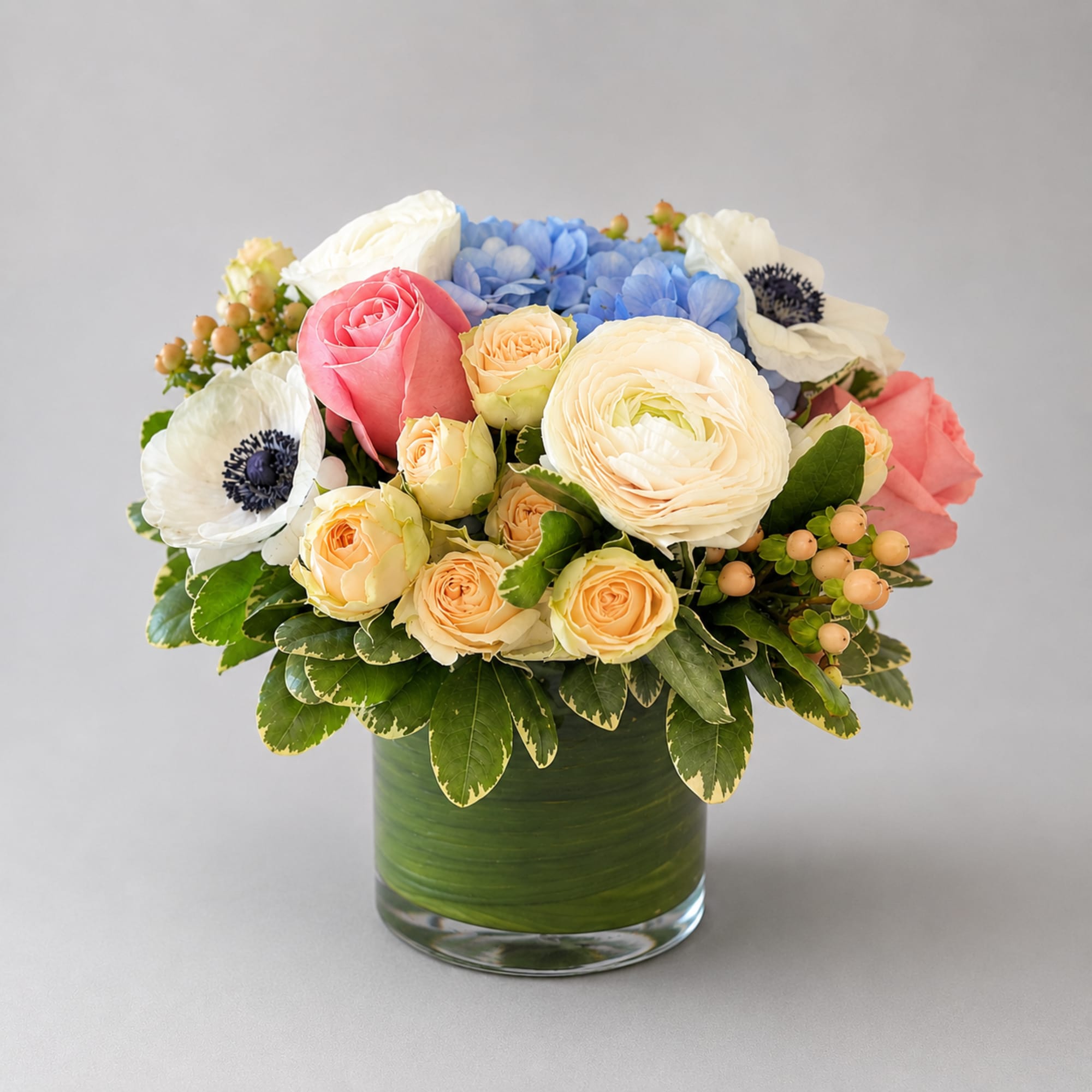 Mixed bouquet of roses, ranunculus, hydrangea, and anemones in a green glass vase