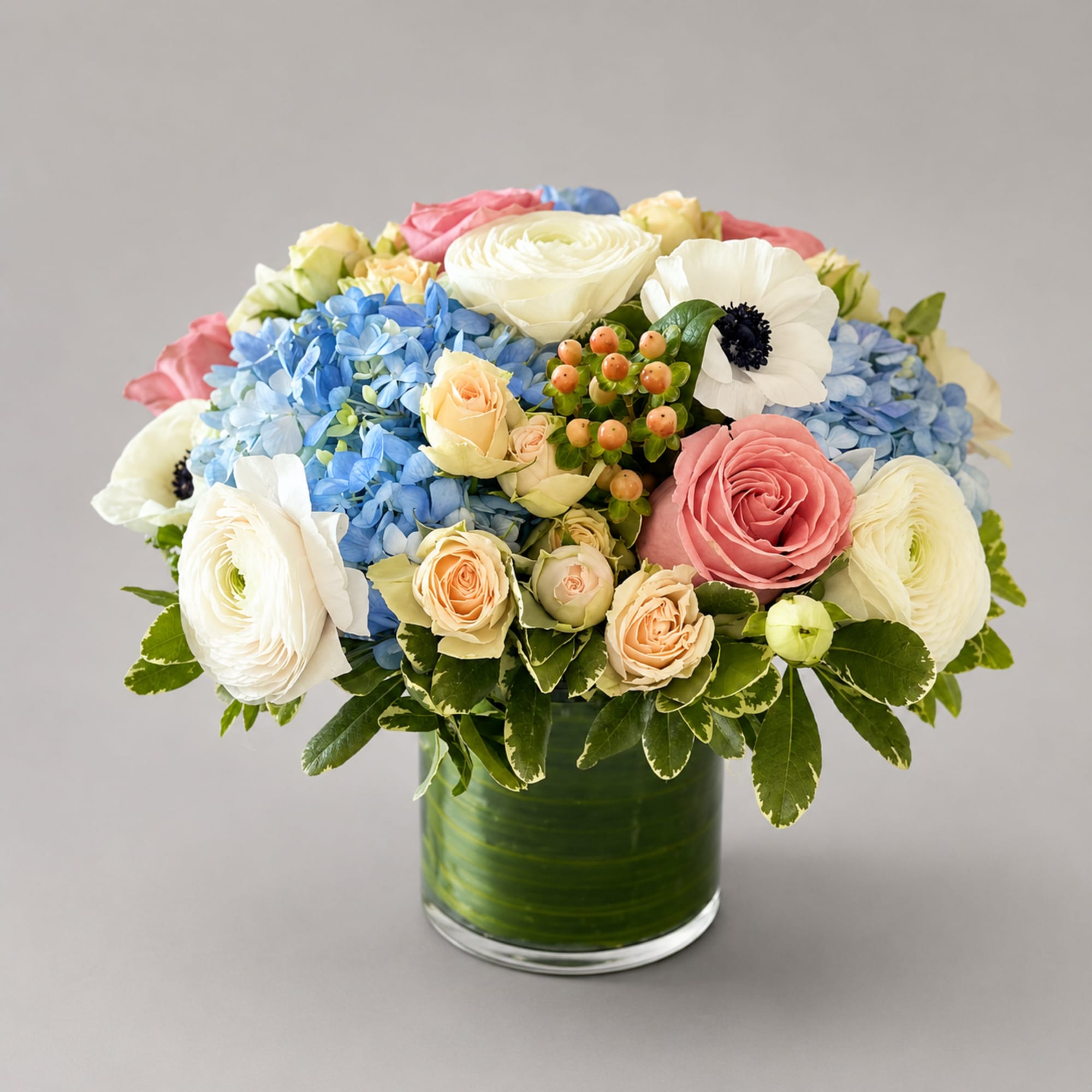 Mixed bouquet of blue hydrangeas, pink and white roses, and white ranunculus in a glass vase