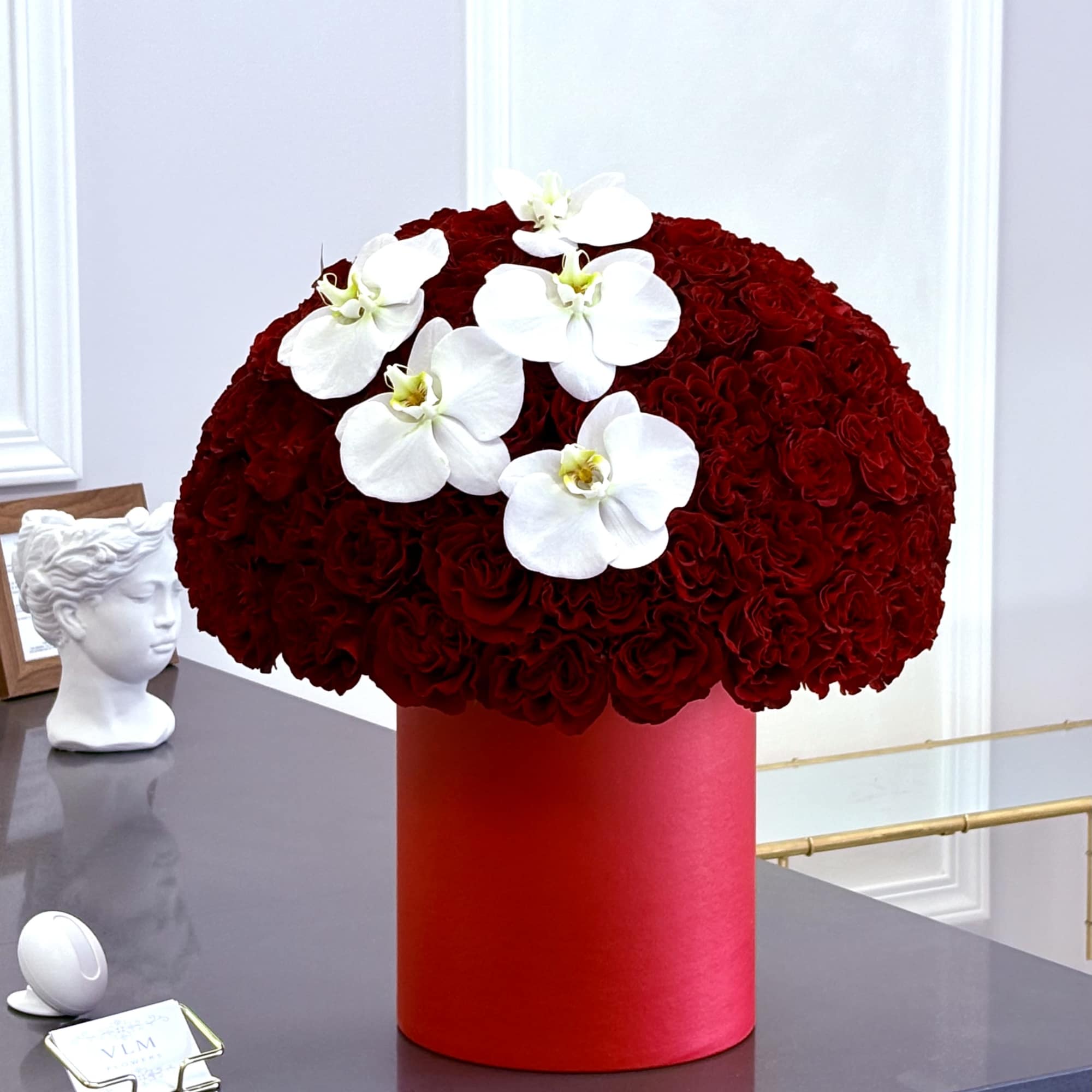 Red rose arrangement in a red cylindrical box with white orchids on top