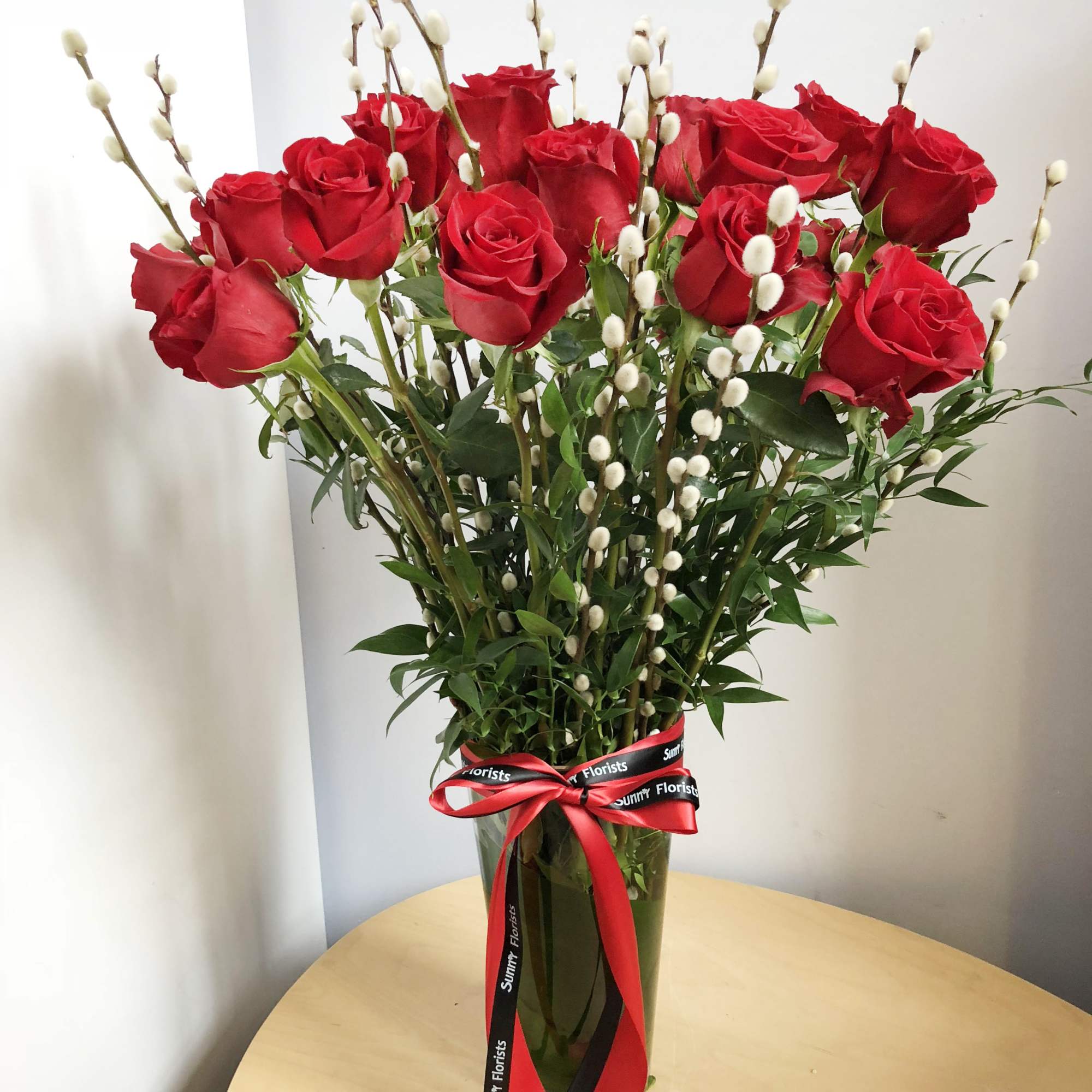 Red roses in a tall glass vase with pussy willow stems and a ribbon bow