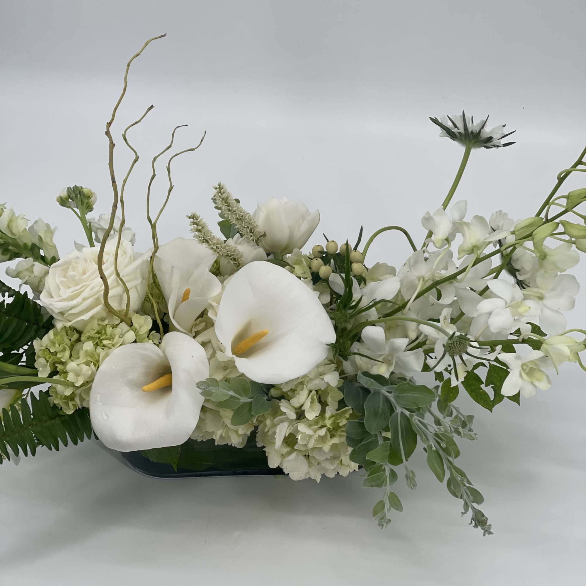 White floral arrangement with calla lilies, roses, and orchids in a low vase