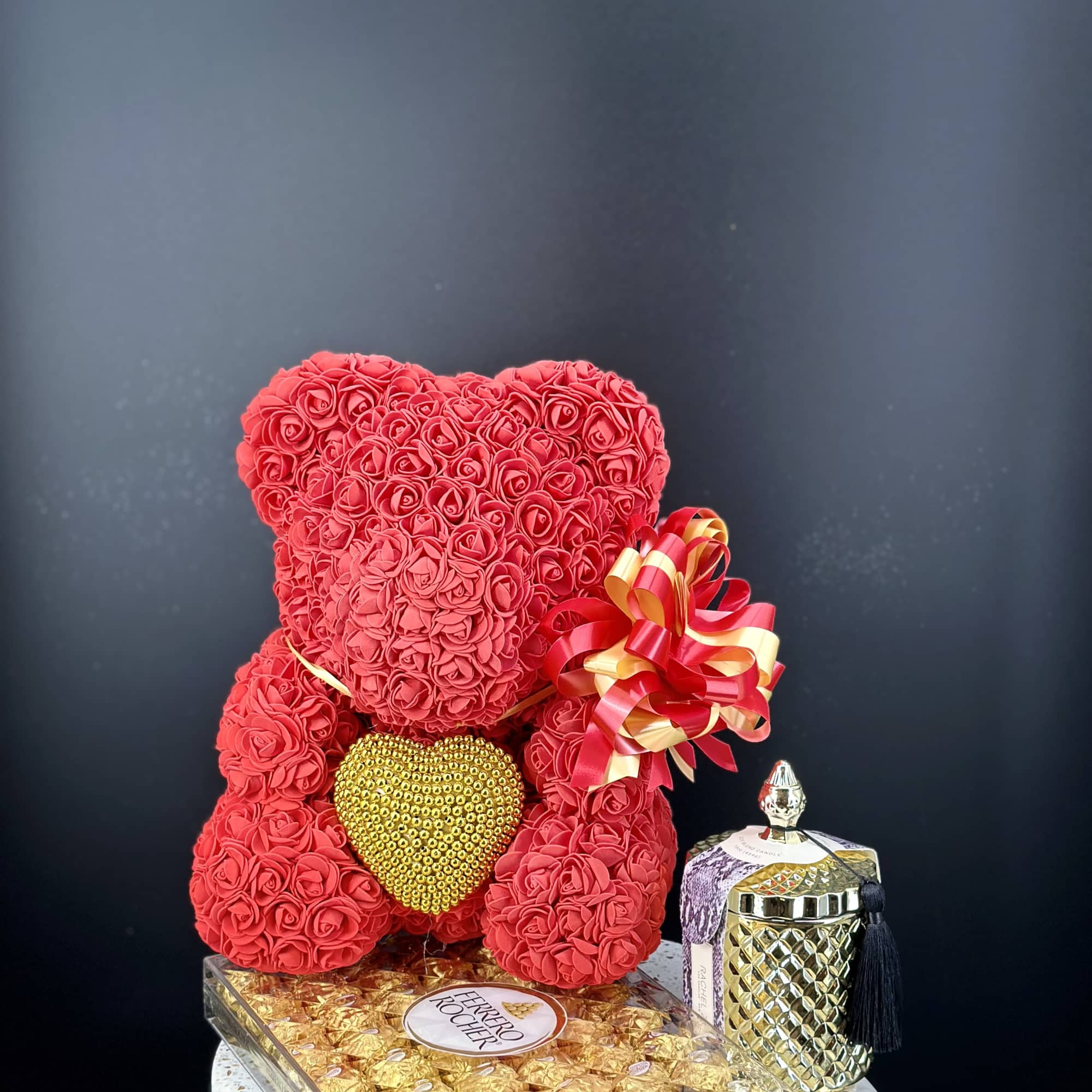 Red rose teddy bear with a gold heart and ribbon bow beside chocolates