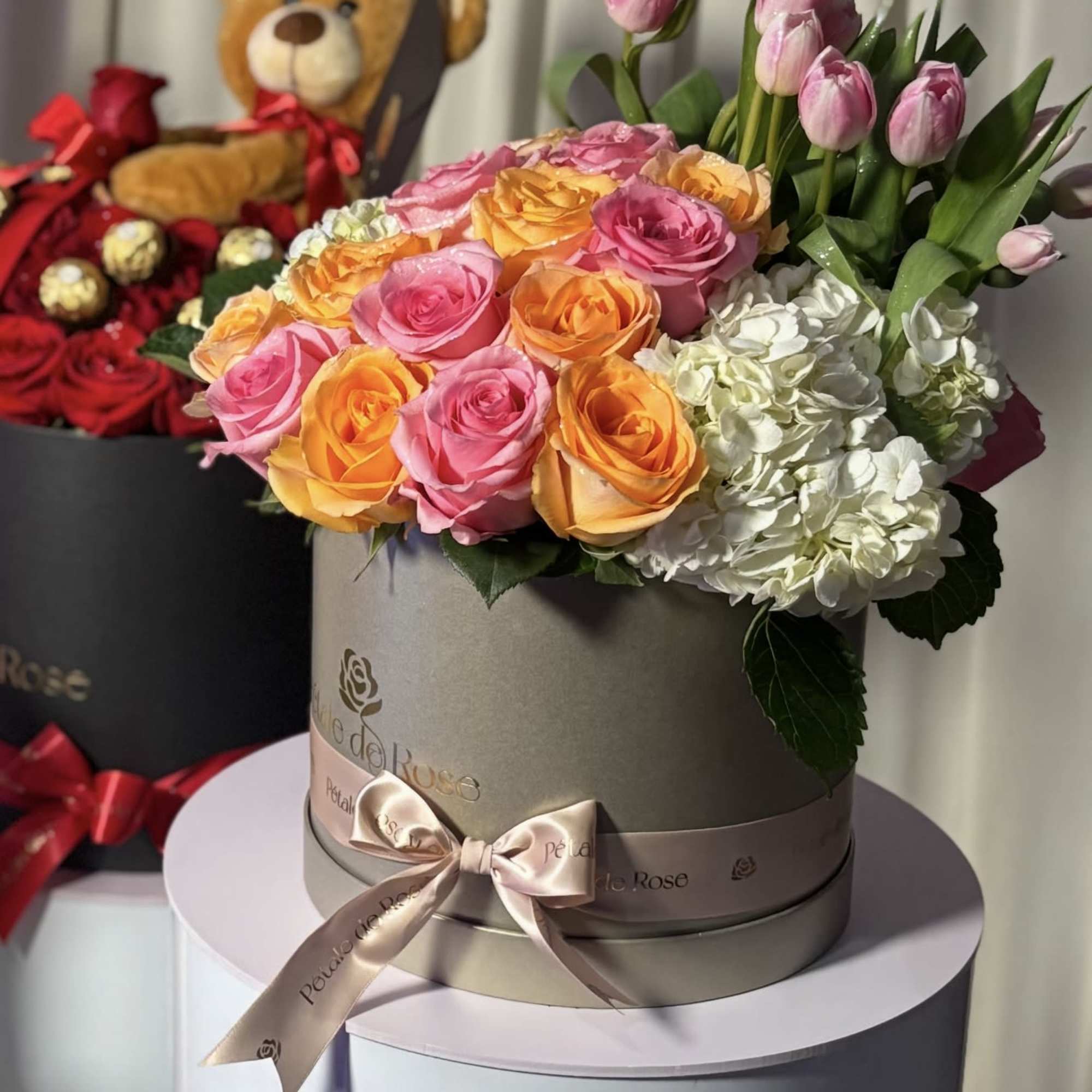 Pink and orange roses with white hydrangeas in a round box, with a teddy bear behind it.