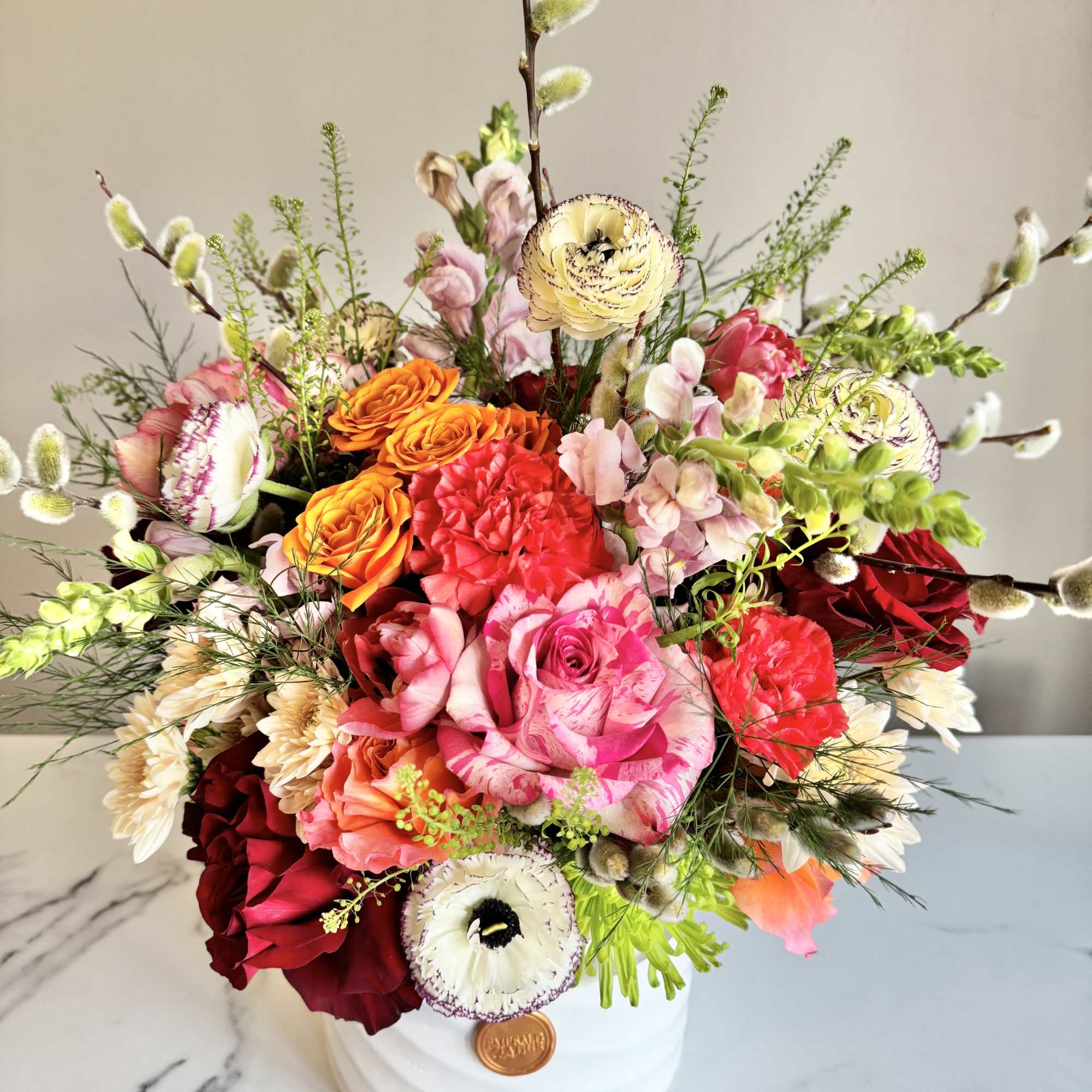 Bright mixed arrangement of pink roses, orange blooms, and white ranunculus in a white ceramic pot.