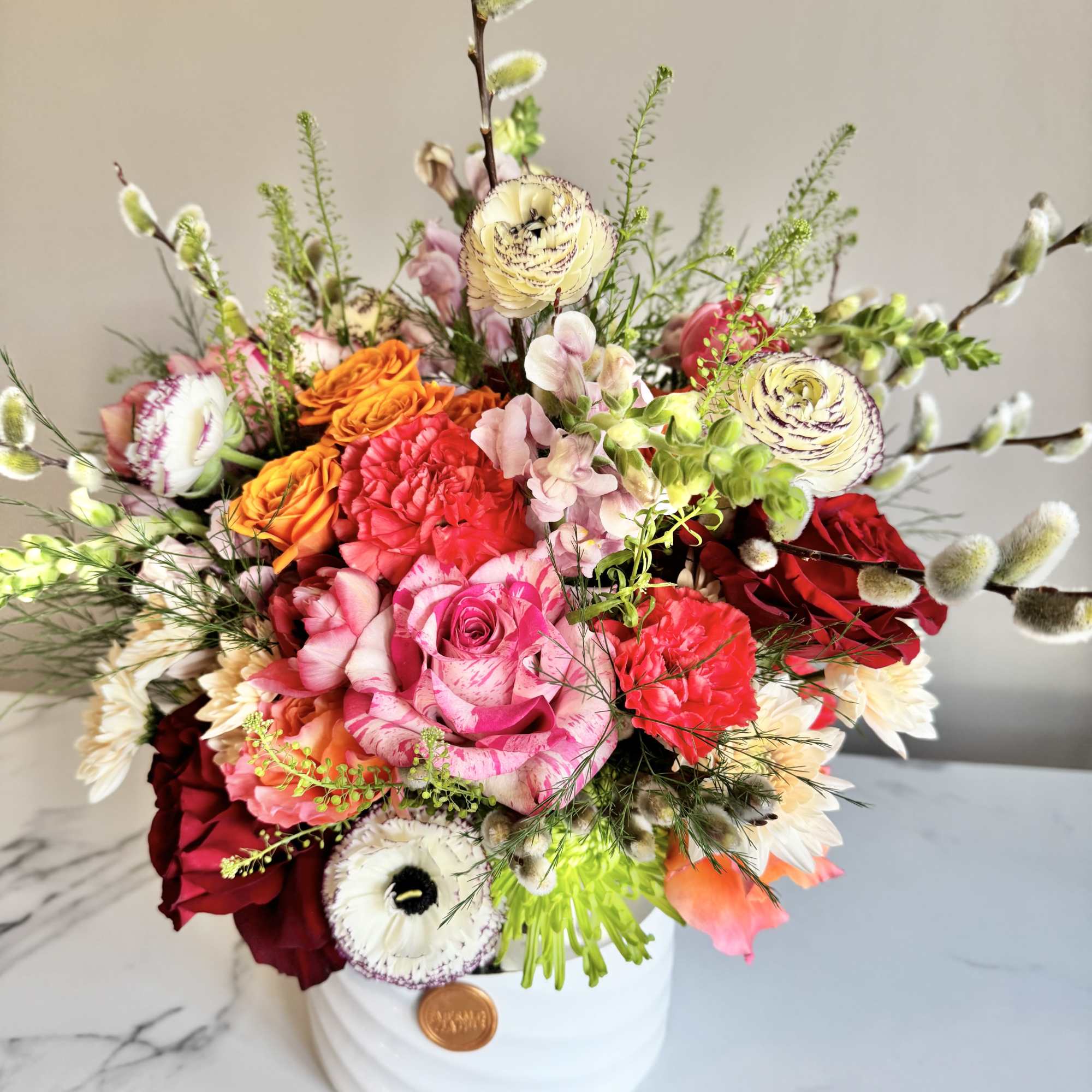 Colorful arrangement of pink, orange, red, and white flowers in a white round vase with tall pussy willow accents.