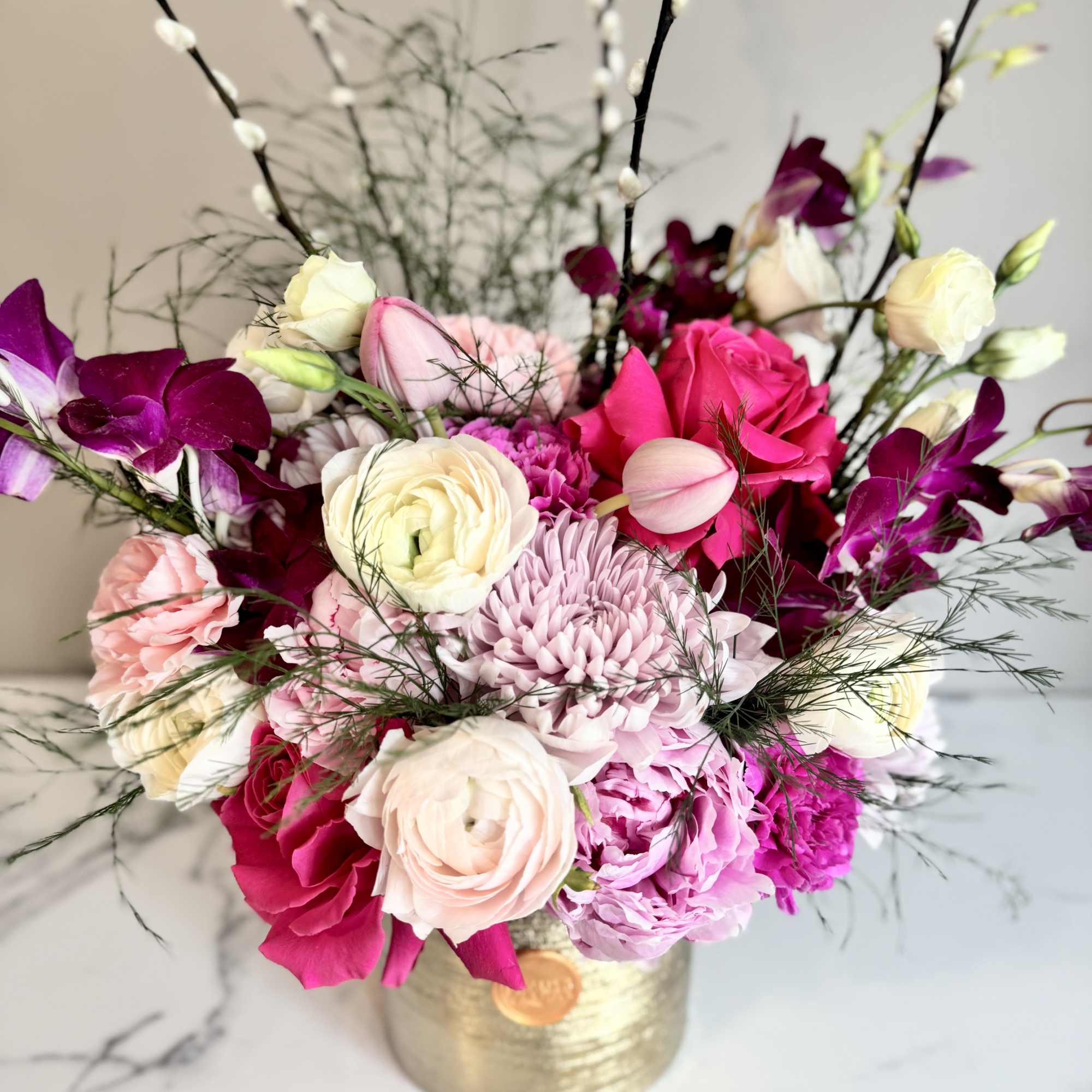 Bright pink, purple, and white mixed flower arrangement in a gold cylinder vase with tall pussy willow stems.