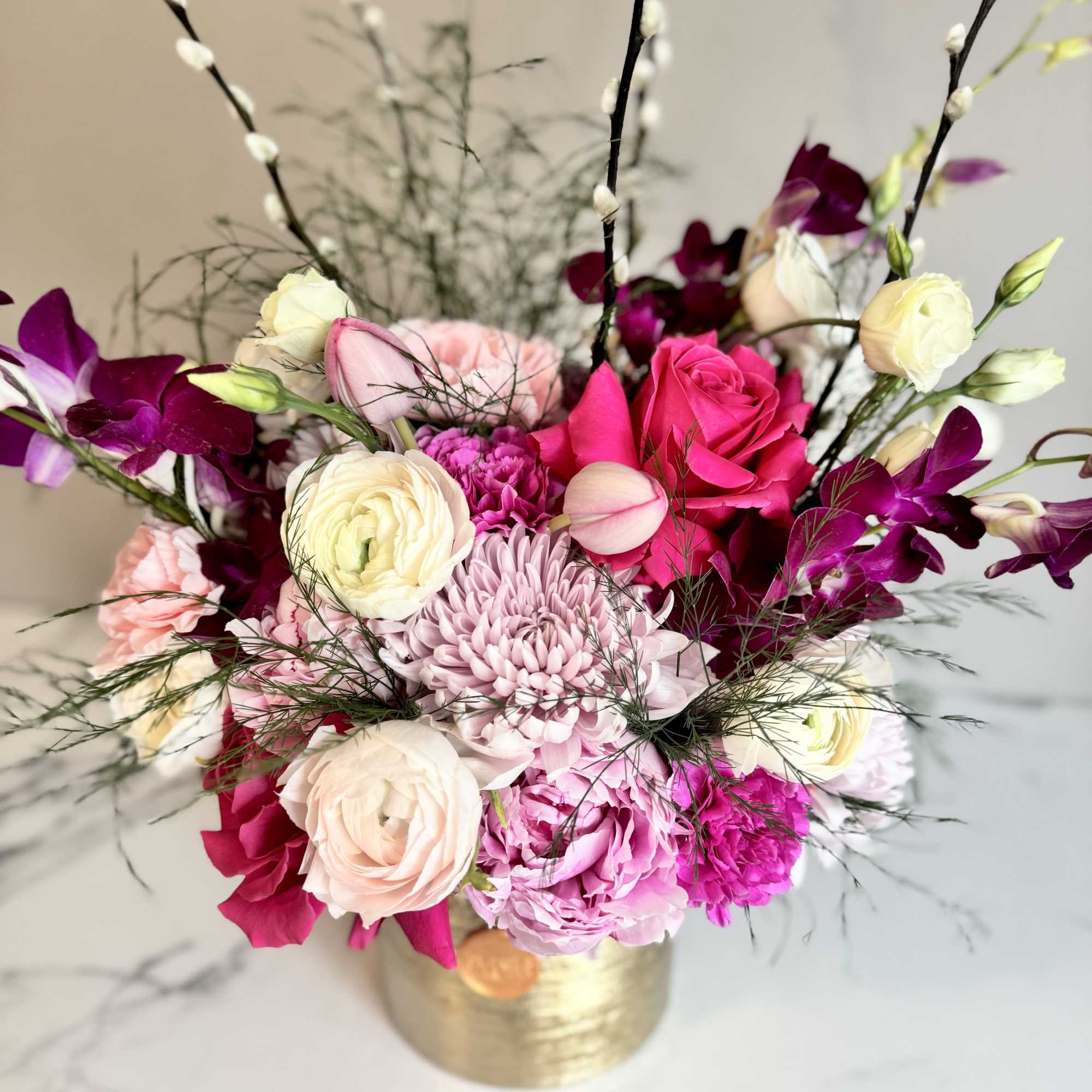 Low arrangement of pink and white roses, ranunculus, orchids and mums in a gold vase with tall pussy willow stems