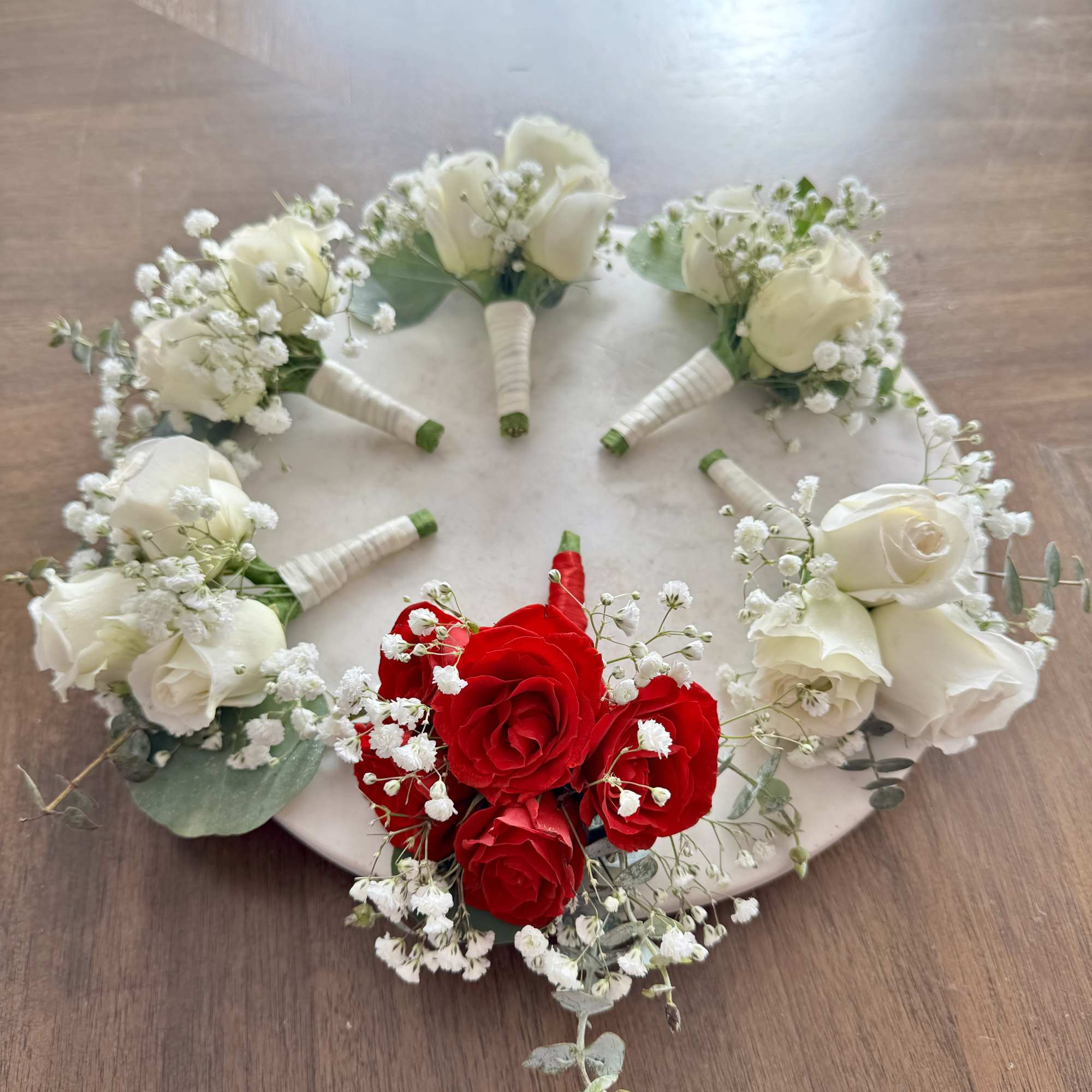 Small rose boutonnieres arranged on a round board, mostly white with one red cluster.