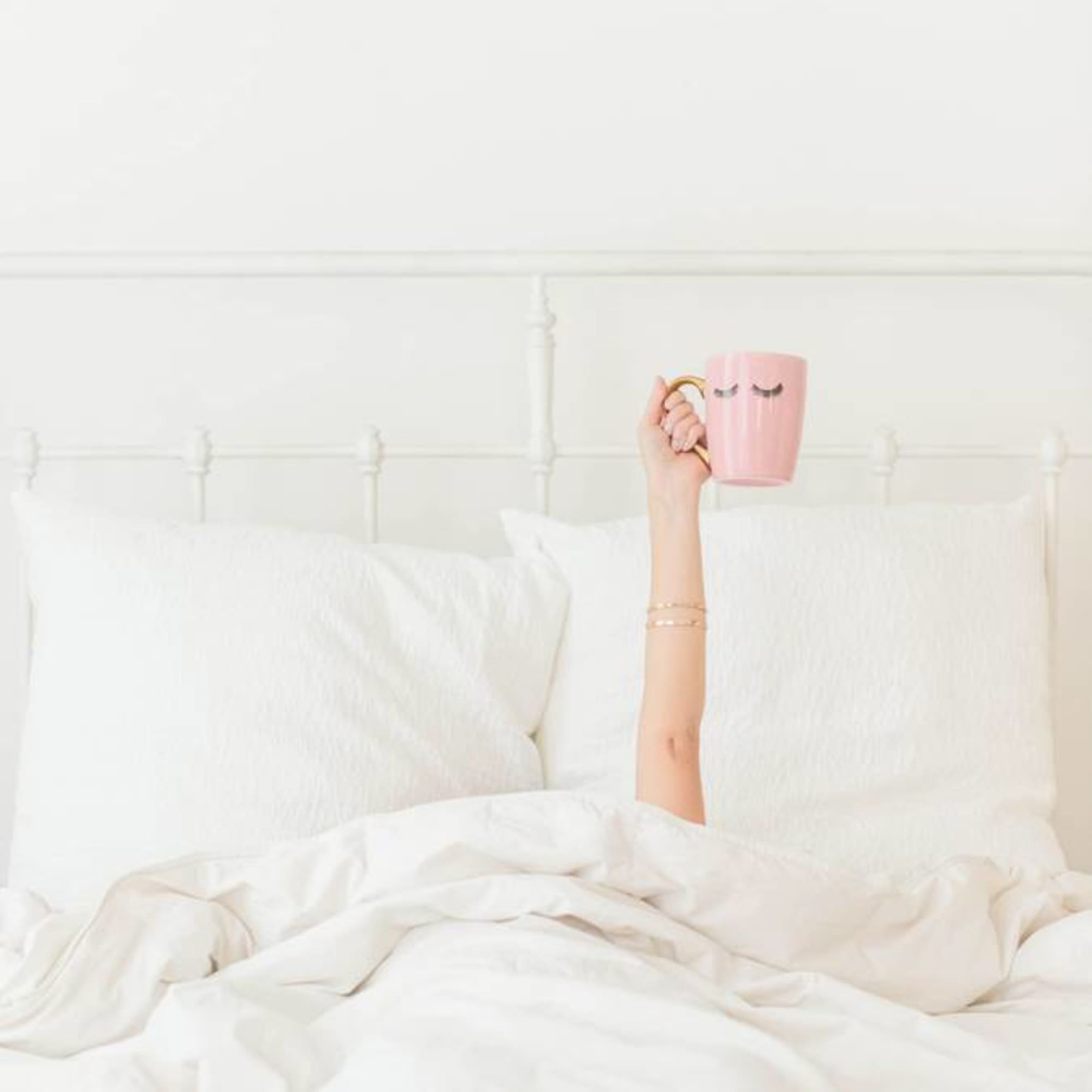 Hand holding a pink mug with closed-eye design above a white bed