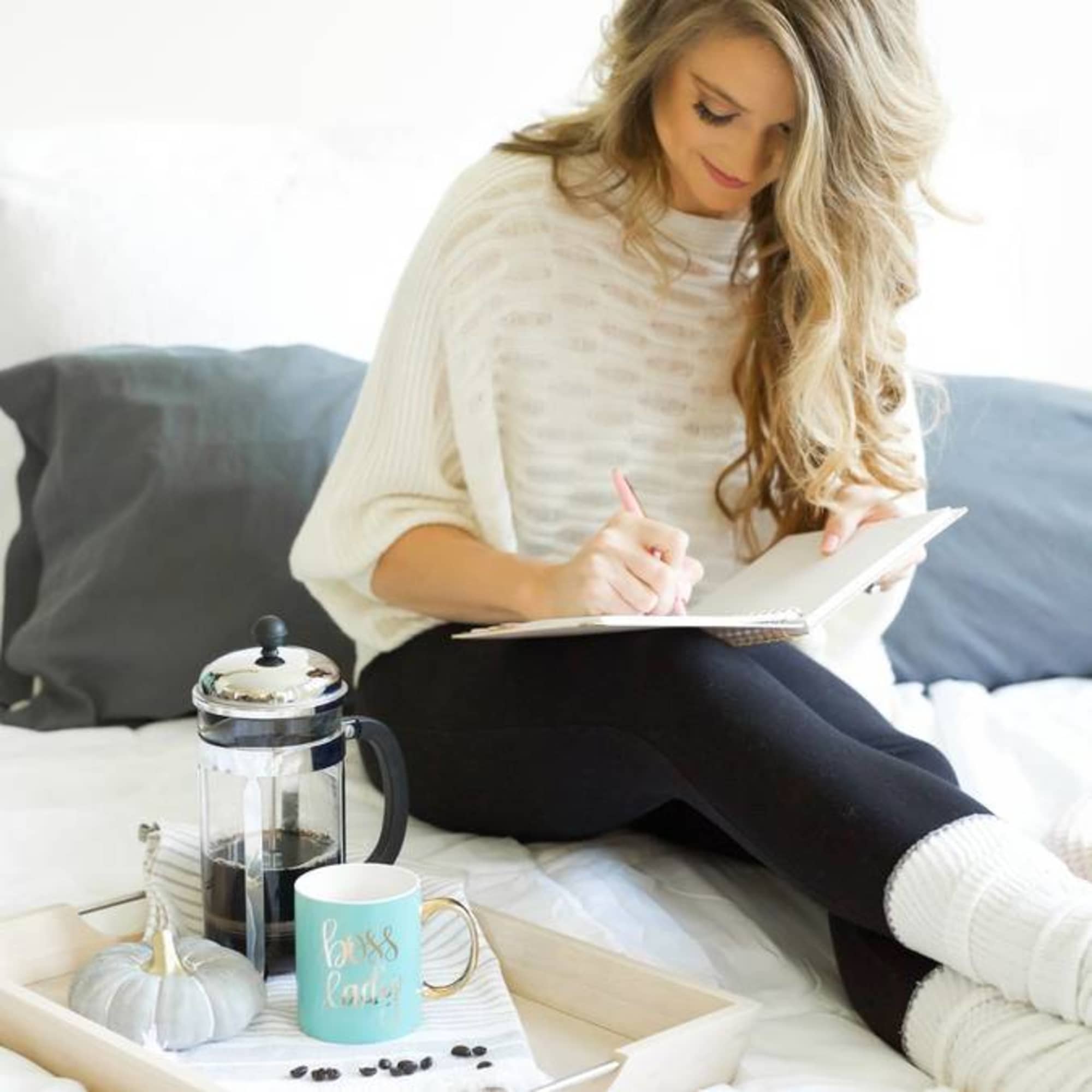 Woman writing in a notebook on a bed beside a coffee tray