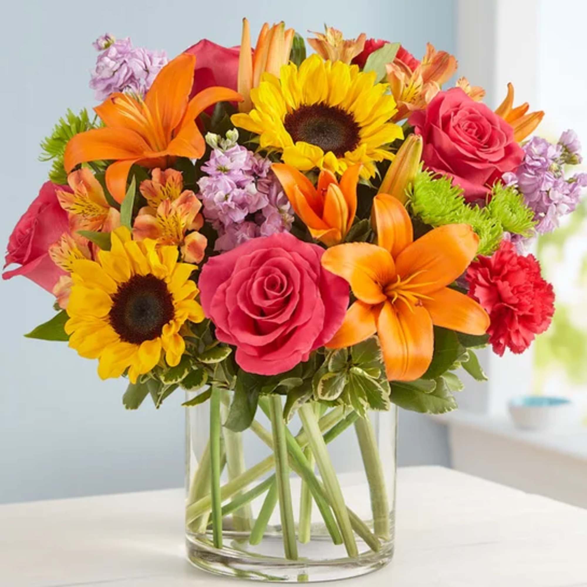 Bright mixed bouquet in a clear glass vase with sunflowers, roses, and orange lilies