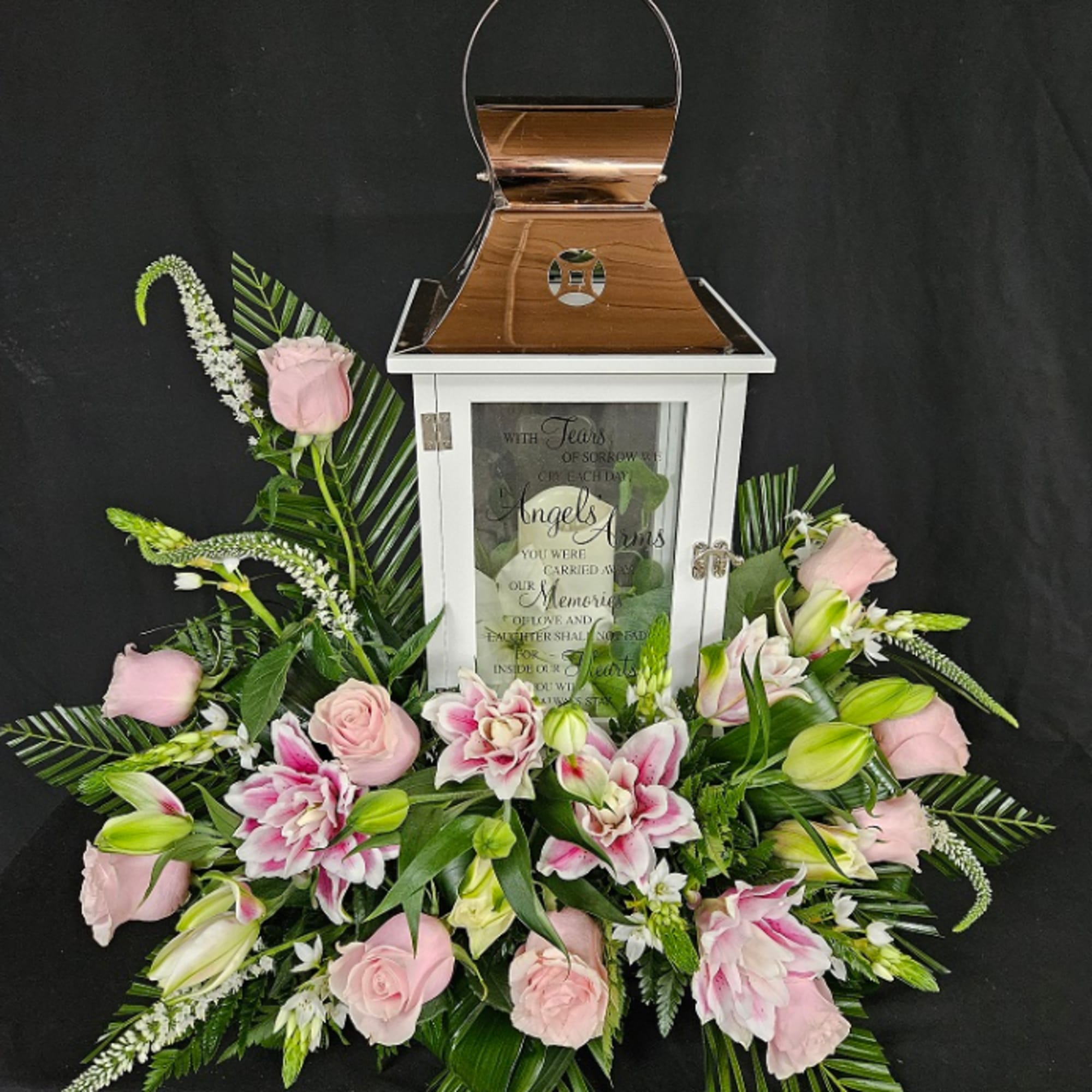 White memorial lantern with copper top surrounded by pink roses and lilies with palm leaves