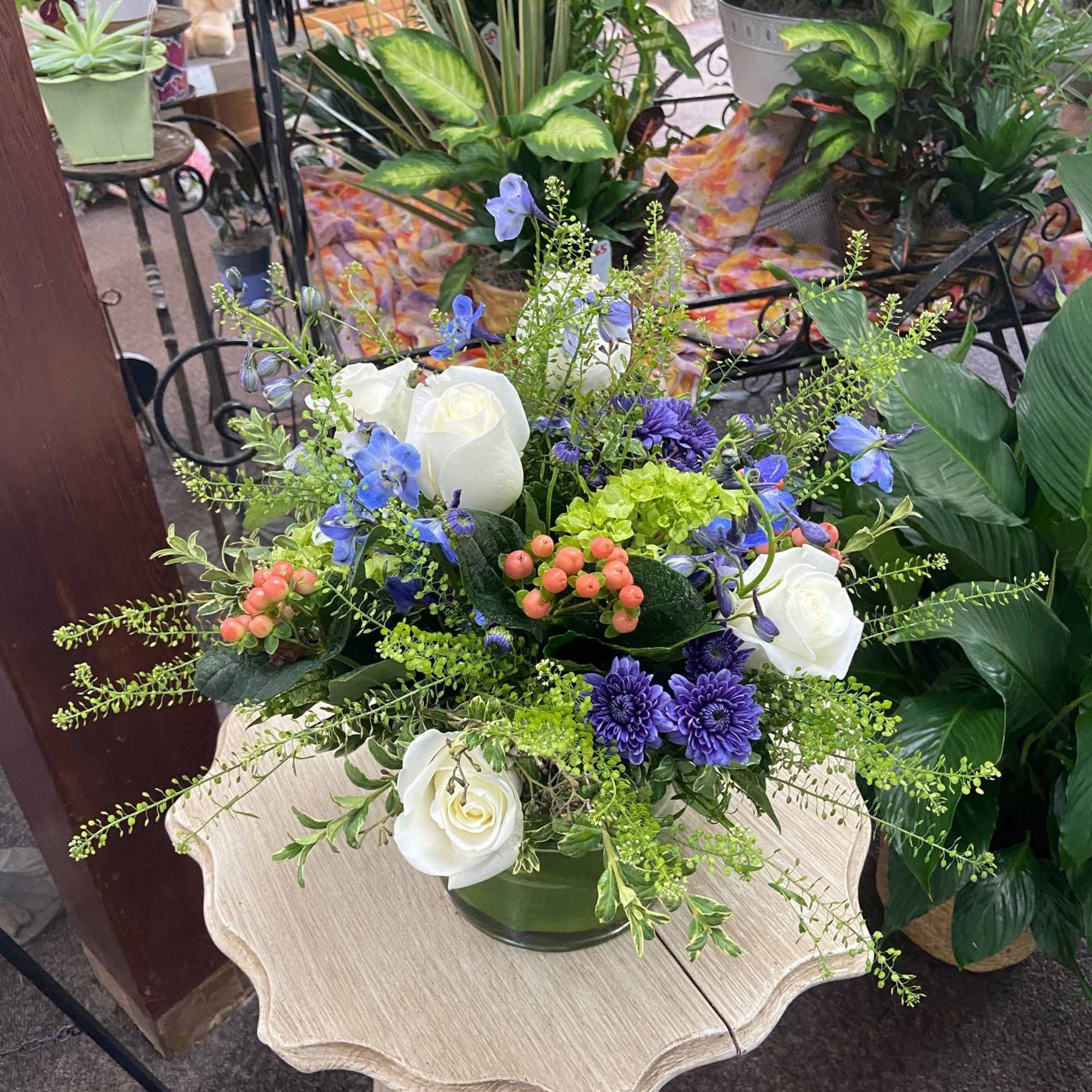 Low arrangement of white roses, purple mums, and blue blooms in a green glass vase.