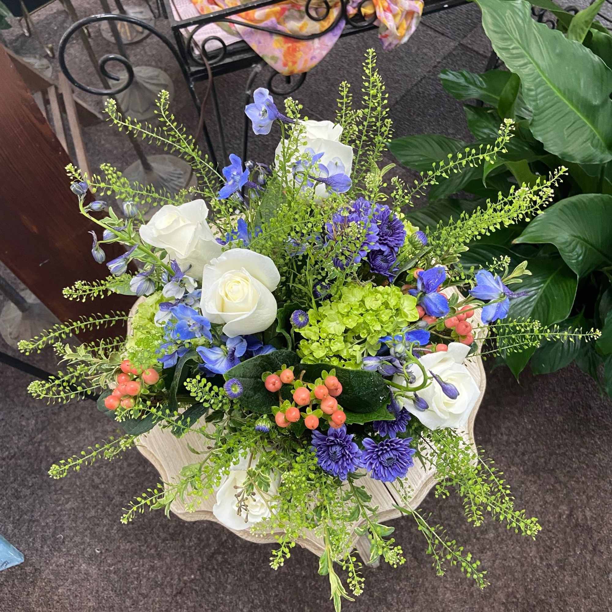 Tall arrangement of white roses, purple blooms, blue flowers, and berries in a cream pedestal container